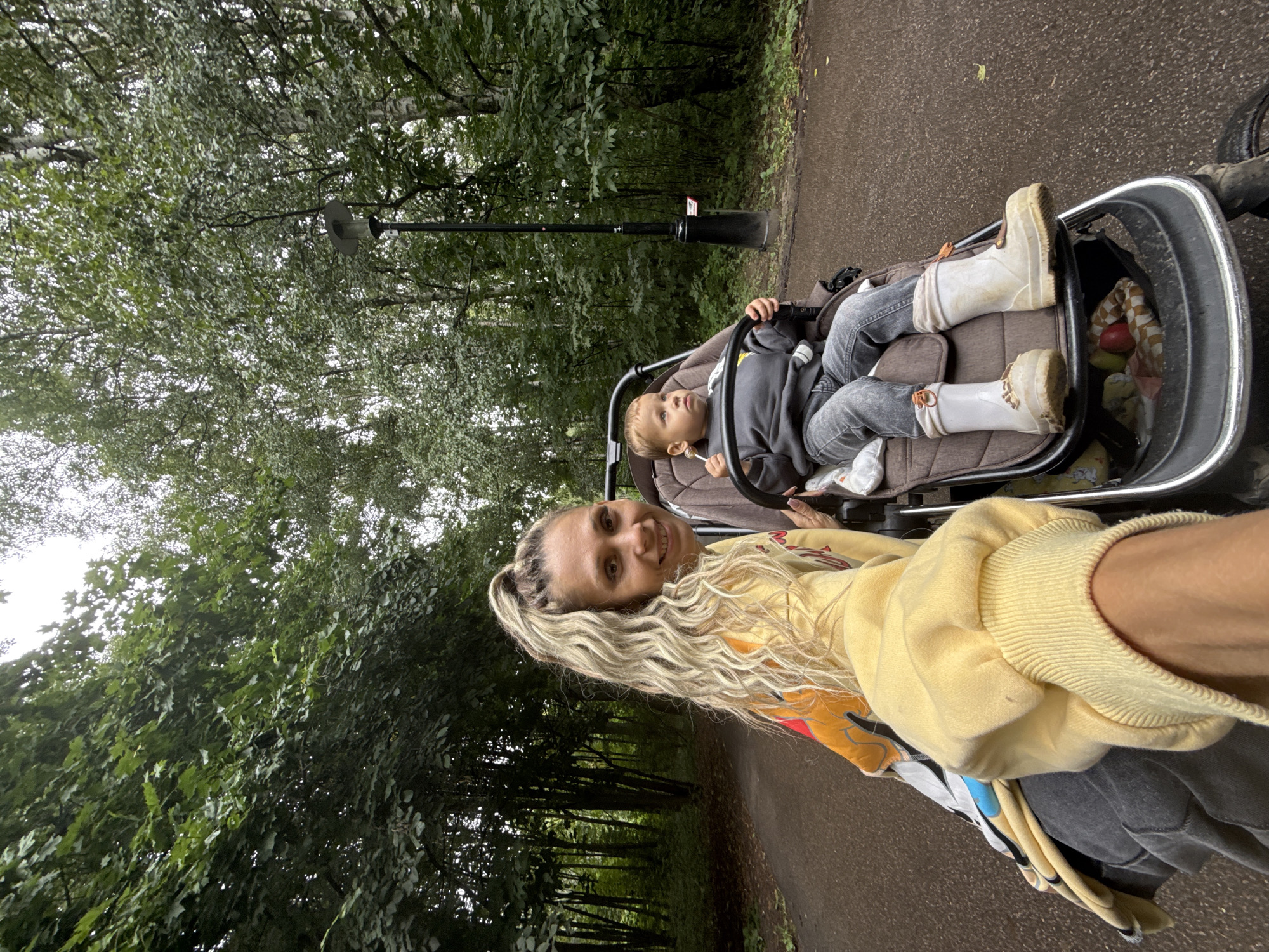 In Rusanovo, Russia, a woman with blonde, wavy hair pulled into a high ponytail is captured in a selfie, smiling at the camera. She is wearing a yellow hoodie with colorful graphic designs on the sleeve and a grey sweatshirt underneath. Beside her, a young child sits in a brown stroller, wearing a grey sweatshirt and jeans. The child holds a lollipop and is looking to the side. The stroller has white boots on the child's feet. The scene takes place on a paved pathway in a park, with lush green trees and foliage on either side. A lamppost is visible in the background. The overall atmosphere suggests an outdoor outing in a natural setting.