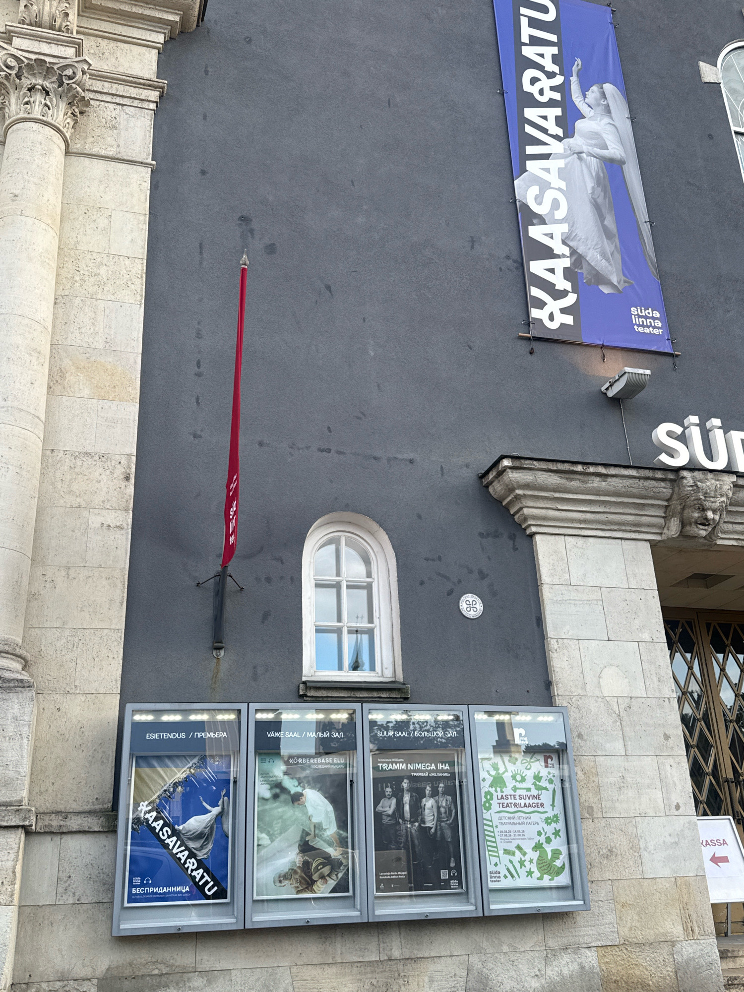 The image shows the exterior of a building in Tallinn, Estonia, likely a theater, with posters displayed on its wall. In the upper part of the building, a large blue banner with white text reads "KAASAVARATU" and features an image of a woman in a white dress. Below this banner, there is a smaller red flag attached to the wall. The wall of the building is dark gray. On the lower part of the building, several framed posters are displayed in illuminated boxes. One poster, with the text "KAASAVARATU" and "ESIENTENDUS / ПРЕМЬЕРА", shows a woman in a white dress against a blue background. Adjacent to it, another poster displays a scene with people in green and white tones. Further to the right, a poster for "LASTE SUVE TEATRILAAGER" (Children's Summer Theater Camp) features green illustrations of children's activities and a dragon. The building's facade is made of light-colored stone blocks, and a classical column is visible on the left side. A sign with the word "KASSA" and an arrow pointing left is visible on the right side of the image. A portion of a door with a metal grate is also visible on the right. The overall scene depicts an outdoor view of a cultural institution.