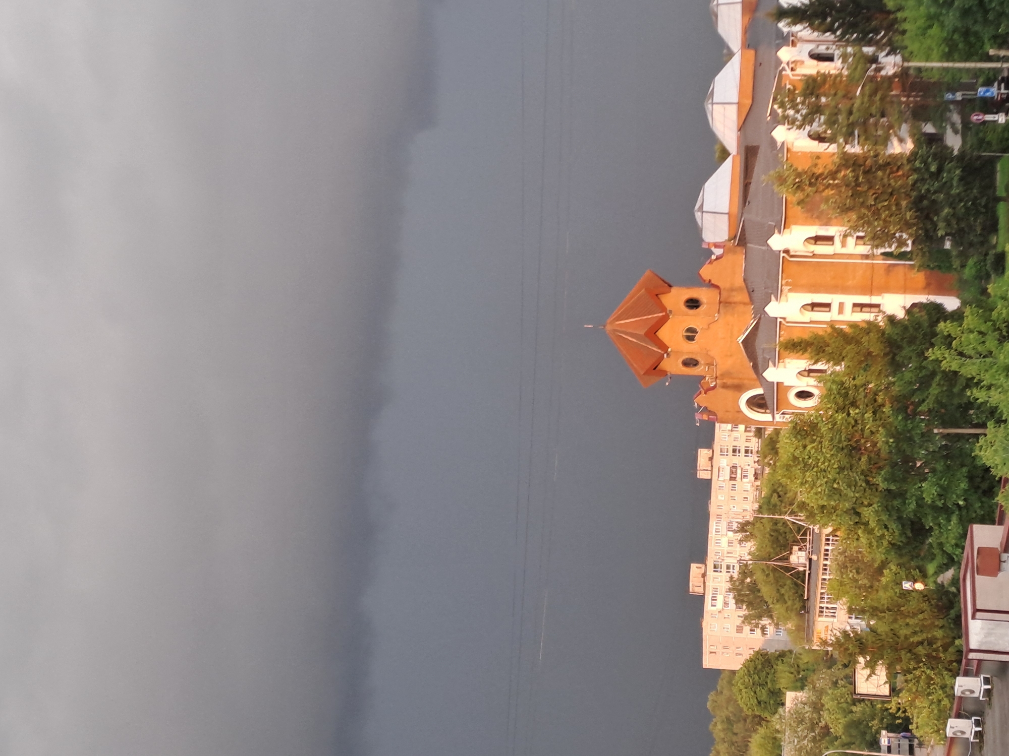 The image depicts a cityscape under a dark, overcast sky, suggestive of impending rain or a storm. In the foreground, a multi-story building with an orange facade and white architectural details is visible. This building features several arched windows and a distinctive turret with a red, hexagonal roof. Several trees, lush and green, surround this building, with some appearing to be evergreen.
Behind this structure, a tall, pale apartment building with numerous windows stretches across the middle ground. This building has a tiered facade and appears to be a residential complex. Further in the background, the sky is dominated by heavy, dark grey clouds, creating a dramatic and somber atmosphere. Faint power lines are visible against the clouds in the distance.
At the very bottom of the image, parts of what appear to be rooftop air conditioning units or similar mechanical equipment are visible on a lower building, along with the edge of a white wall. The scene is set in the city of Elektrostal, Russia, as indicated by the building architecture and urban environment. No people are clearly visible in the frame. The overall impression is one of a city bracing for inclement weather.