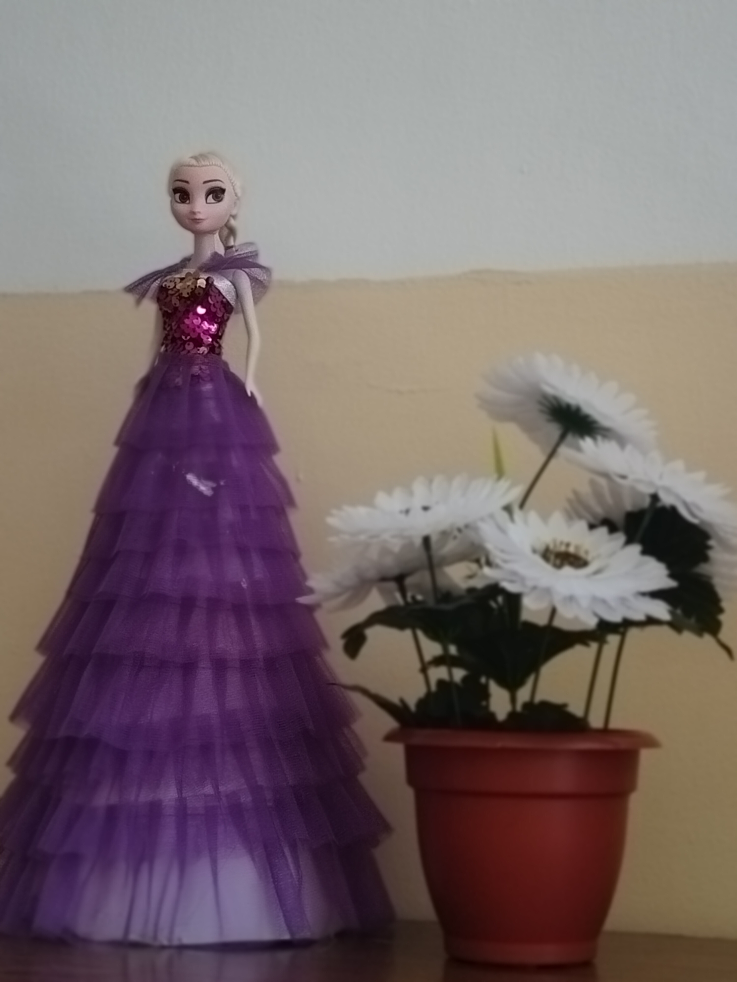 A doll, resembling Elsa from the movie Frozen, stands to the left of the frame. It is wearing a layered purple gown with a sequined bodice. To the right of the doll, a potted plant with several white artificial flowers is displayed. The plant is in a brown pot and sits on a wooden surface. The background consists of a light-colored wall. The scene appears to be indoors, possibly in a domestic setting. There is no indication of the location being in the city of Көлкент, Kazakhstan.