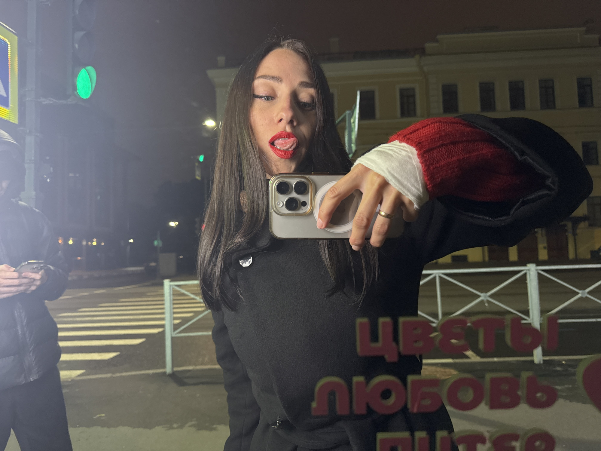 A woman stands on a city street at night, holding a smartphone and taking a selfie. She has dark hair and is wearing red lipstick. She is sticking her tongue out. She is wearing a dark coat and a red and white knit sleeve.
In the background, a pedestrian looks at their phone. A green traffic light is visible. A crosswalk is visible and a building is behind the woman, and the text "ЦВЕТЫ ЛЮБОВЬ ПИТЕР" is printed on the glass she is standing in front of.
The text translates to "Flowers Love Peter" which is a common name for Saint Petersburg, Russia. Therefore, the location is in Saint Petersburg, Russia.