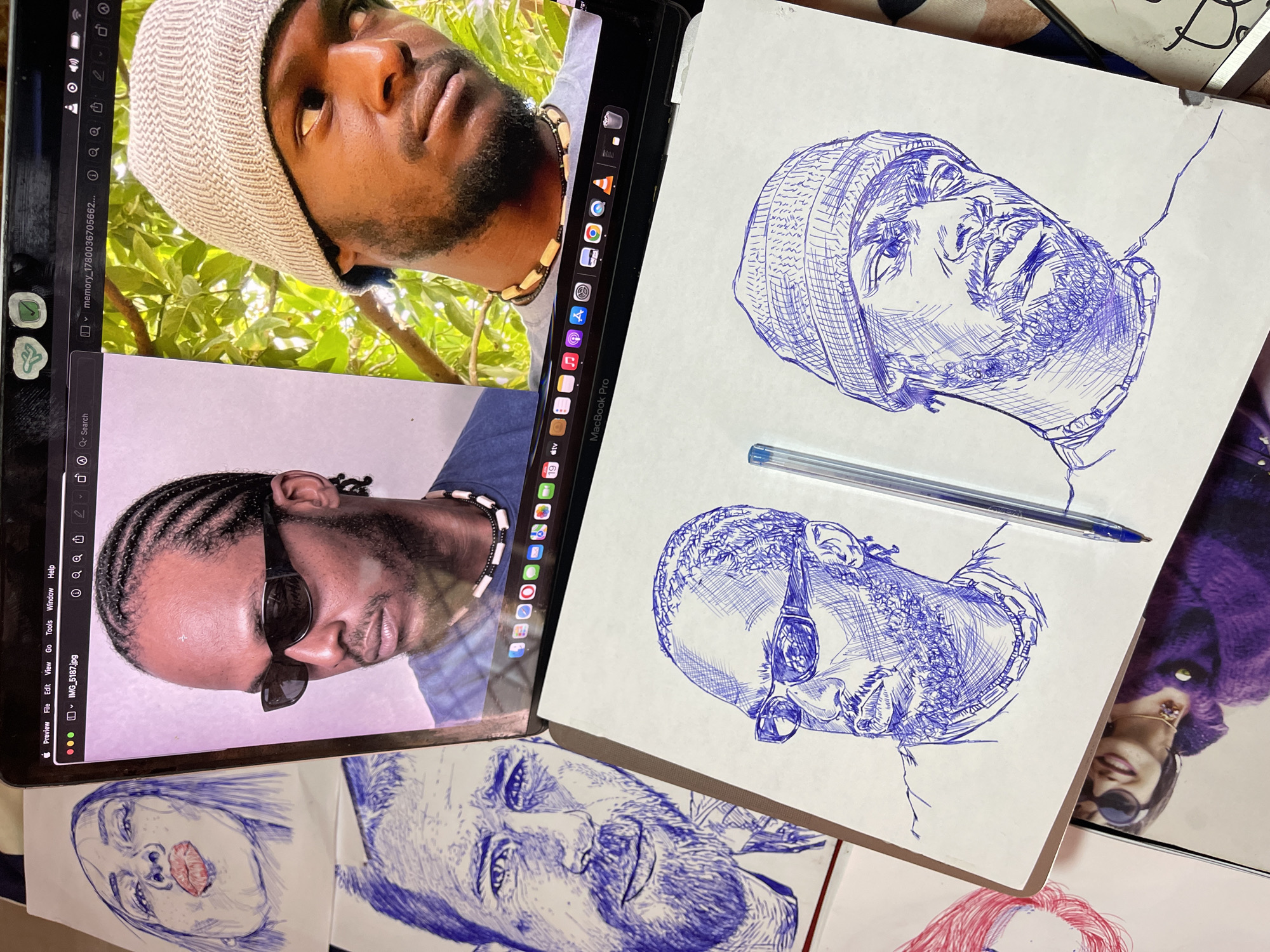 The media file displays a close-up, overhead shot of a workspace featuring an open laptop and several sketches. The laptop screen shows two candid photographs of a Black man. In the upper image, he is wearing a beige knit cap and looking upwards, with green foliage in the background. The lower image shows him wearing dark sunglasses and a blue shirt, with his hair styled in braids. A bead necklace is visible around his neck.
On the right side of the frame, a sketchbook lies open, showcasing two blue ink sketches of the same man, also wearing a beanie and sunglasses in one sketch, and a beanie in the other. A blue ballpoint pen rests across the sketchbook pages. To the right of the sketchbook, part of a purple fuzzy garment is visible, and below it, another sketch of a woman's face with red lips and black sunglasses can be seen. Further down, a sketch of a man's face with a beard and a sketch of a woman with pink hair are also visible.
The scene appears to be indoors, possibly a creative studio or home office, with the lighting suggesting daytime. The overall mood is artistic and focused, with the sketches and the pen indicating an ongoing creative process. No explicit time of day or weather cues are discernible, nor are there specific location cues beyond the provided context of Jos, Nigeria. The subject's emotions are largely neutral in the photographs and artistic interpretations in the sketches. The laptop screen displays file names like "memory_1780036705662..." and "IMG_5187.jpg." The branding "MacBook Pro" is also visible on the laptop.