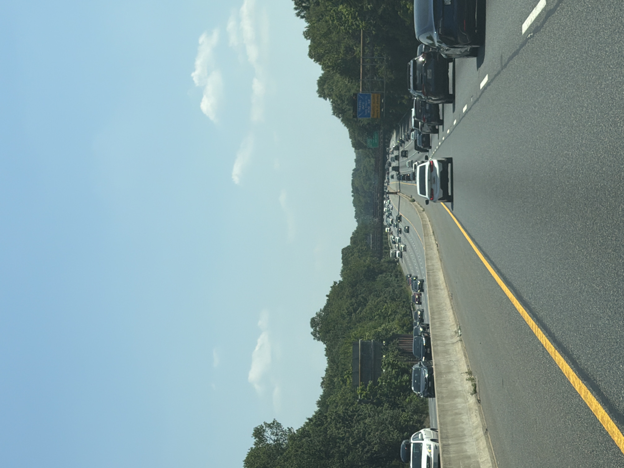 A vertical, eye-level, outdoor, daytime, full shot captures a highway scene in Glenarden, Maryland, United States of America. The sky is a bright, clear blue, with a few wispy white clouds scattered across it. The highway itself is asphalt and has a prominent yellow line down the center of the lane. Several cars are on the highway, driving in the same direction. The cars closest to the camera include a white SUV on the left, a black SUV in the center, and a dark-colored sedan on the right. Further down the highway, more cars are visible, appearing as smaller shapes. Trees line both sides of the highway, forming a dense green wall. In the distance, overpasses and highway signs are visible, indicating the complex nature of the road system. The overall impression is one of a typical, busy highway commute on a sunny day.