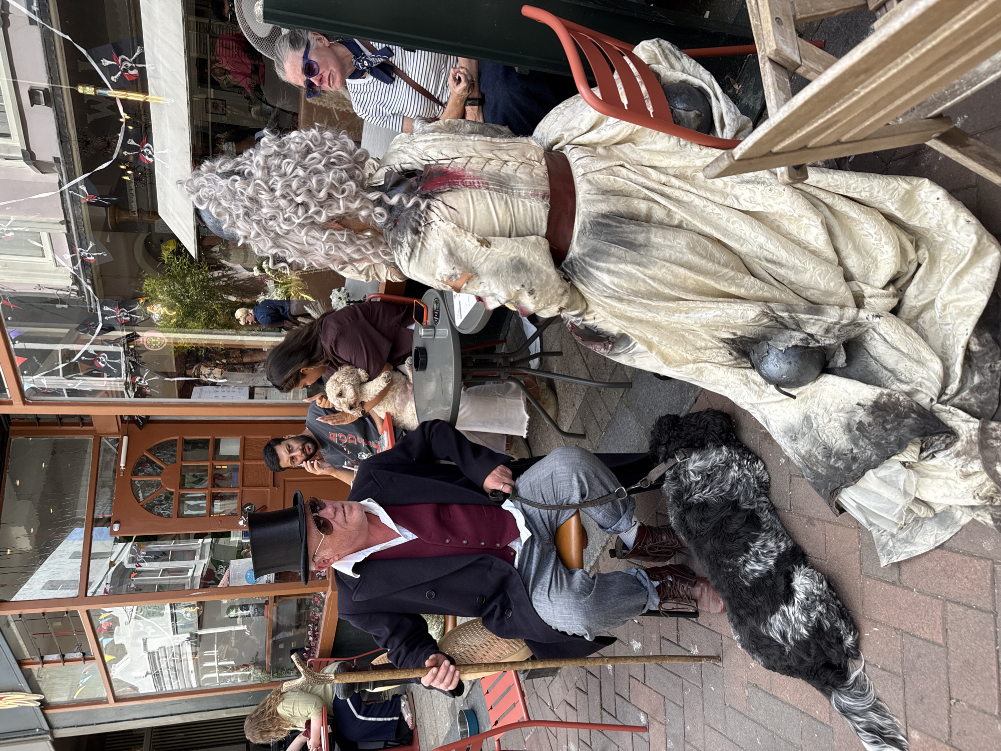 In St Leonards, United Kingdom, outside a building with a glass-fronted cafe or shop, two individuals are seated at tables on a paved outdoor area. On the left, a man wearing a black top hat, sunglasses, a dark coat, a burgundy vest, and grey trousers sits in a chair, holding a walking stick and a dog leash. A black and white shaggy dog lies on the ground near his feet. On the right, a person with elaborate, curly grey hair wears a distressed, pale, long dress with dark markings. This person is seated facing away from the camera, with their back partially to the viewer. A second dog, a light-colored poodle, is visible on a table between the two seated individuals. Other patrons are seated at tables in the background. The building facade is decorated with Halloween-themed pirate flags. The atmosphere suggests a themed event or gathering.