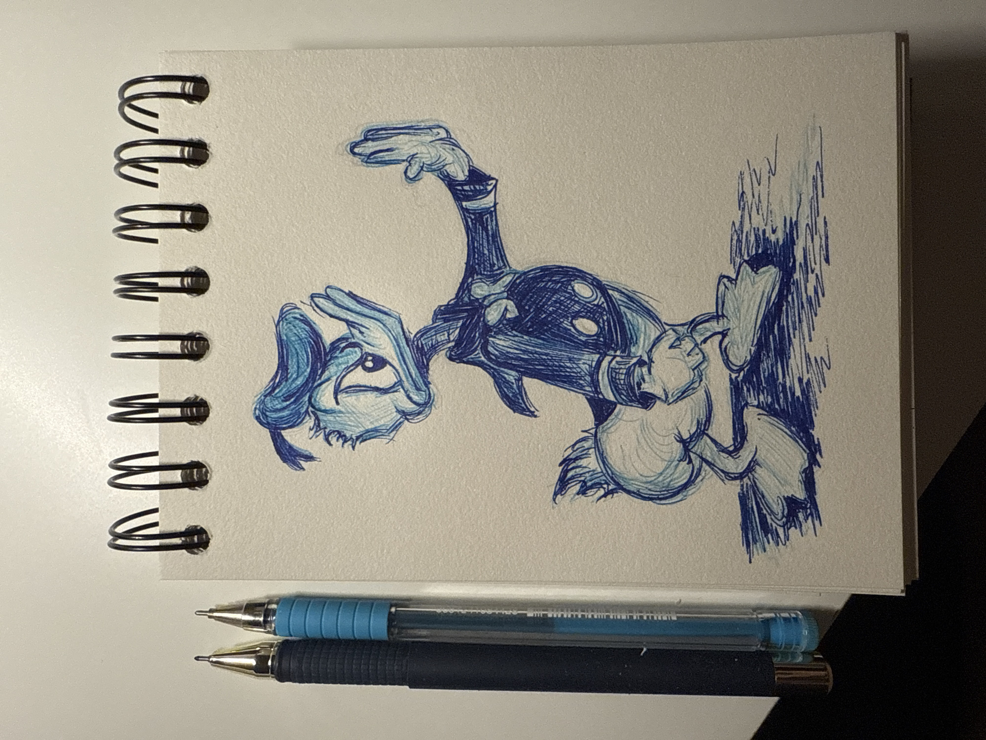A medium, eye-level shot depicts a sketchbook featuring a pen and ink illustration of a cartoon character resembling Donald Duck, lying on its side. The drawing is in blue ink and the character appears to be standing on a wall. Below the sketchbook, two pens are placed horizontally; one with a light blue body and one with a dark blue body. The sketchbook sits on a white surface with darker, shadowed areas towards the right of the frame. The sketchbook is spiral-bound. No people are visible. There is no indication that the location is Sofia, Bulgaria.