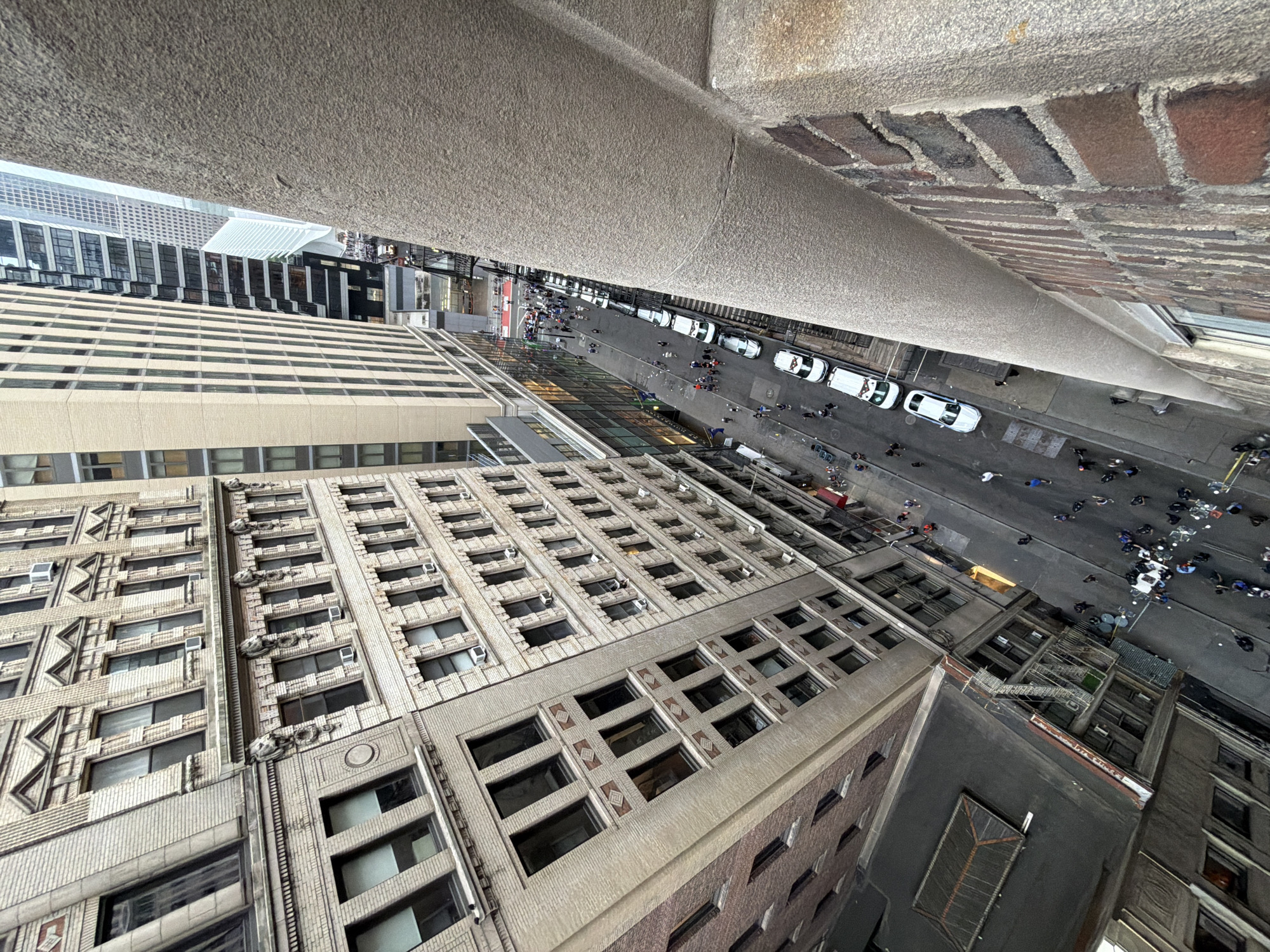 A high-angle, exterior, medium shot captures a narrow city street in New York City, USA, lined with tall buildings on both sides. The street is mostly empty, with a few cars parked along the side and a small group of people walking. The buildings are a mix of old and new architecture, with some featuring ornate brickwork and others displaying modern glass facades. The sky is overcast, casting a muted light over the scene. On the right side of the frame, a rough, textured concrete wall juts out, obscuring a portion of the view. The overall impression is one of urban density and a quiet, overcast day.
