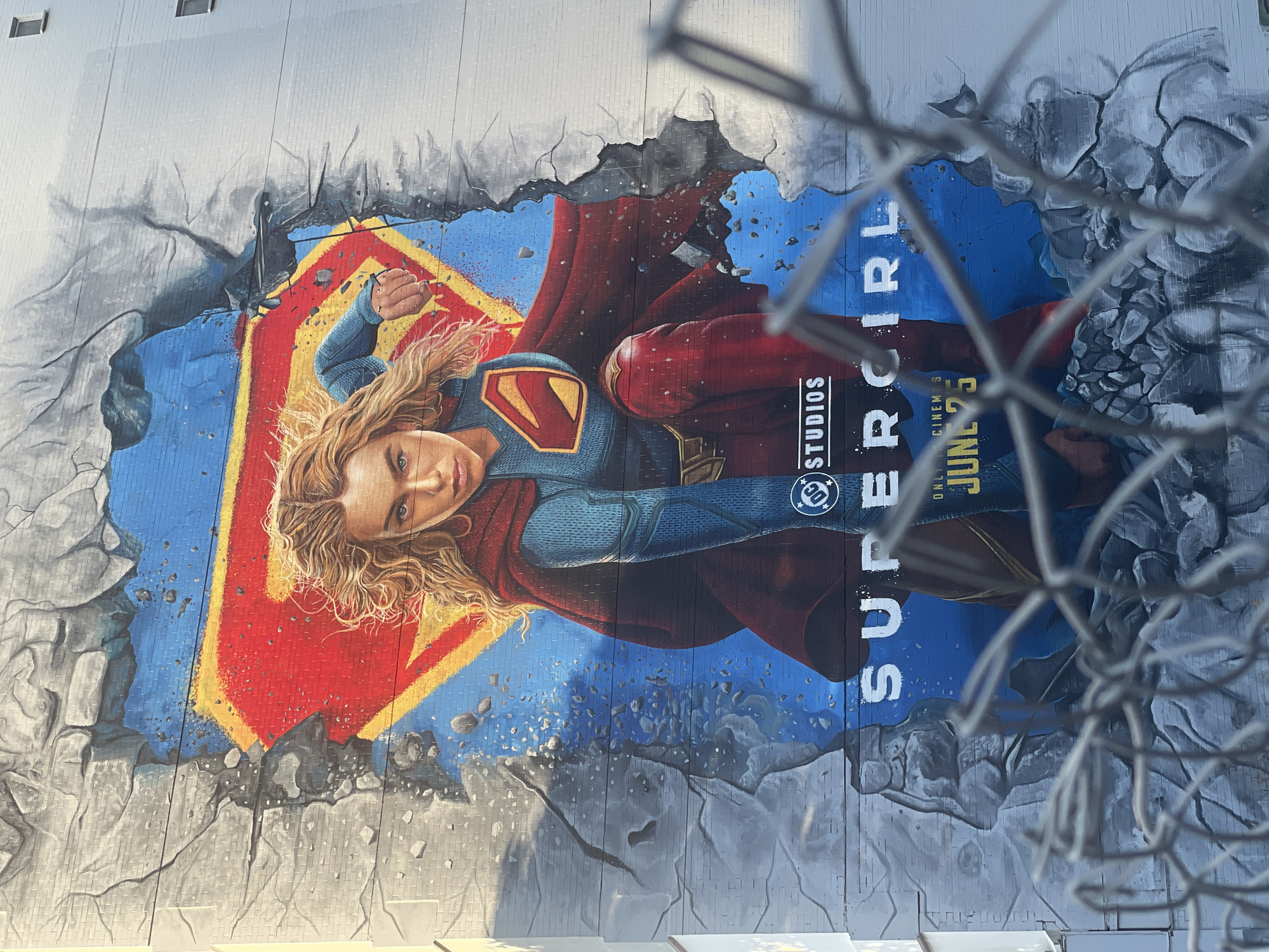 A large mural of Supergirl is painted on the side of a building. Supergirl is depicted in her classic costume with a red cape, blue and red suit, and the iconic "S" shield. She is positioned dynamically, with one knee bent and her arms outstretched, as if bursting through a brick wall. The mural uses vibrant colors, with a bright blue background and a large red and yellow "S" shield behind Supergirl. The illusion of a crumbling brick wall is rendered in grayscale, creating a dramatic contrast with the colorful superhero. Text on the mural reads "DC STUDIOS" and "SUPERGIRL" with "JUNE 25" underneath, indicating it is a promotional artwork for the movie. The mural is viewed through a chain-link fence, which partially obscures the lower portion of the artwork. The building appears to be a multi-story structure, and the mural covers a significant portion of its facade. This mural is located in the Fortitude Valley area of Brisbane, Australia, specifically on Ann Street.