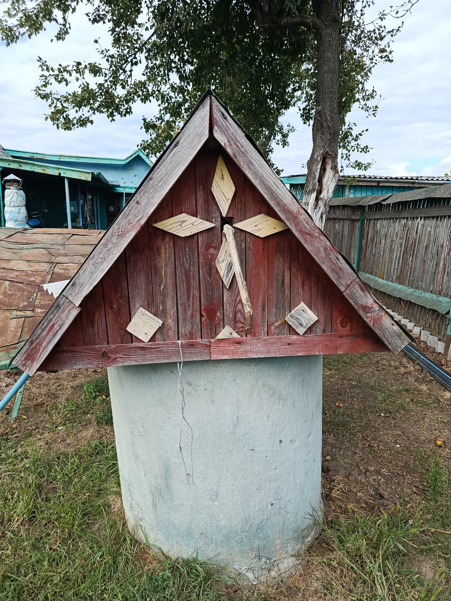 A well with a triangular wooden roof, painted red and adorned with diamond-shaped wooden accents and a central cross made of planks, stands in a grassy area. The well's concrete base is weathered and shows some signs of wear. In the background, a rustic wooden fence, a tree with a visible hollow, and parts of buildings with corrugated metal and painted siding are visible. The sky is overcast with some clouds. The setting appears to be rural, possibly a yard or garden.