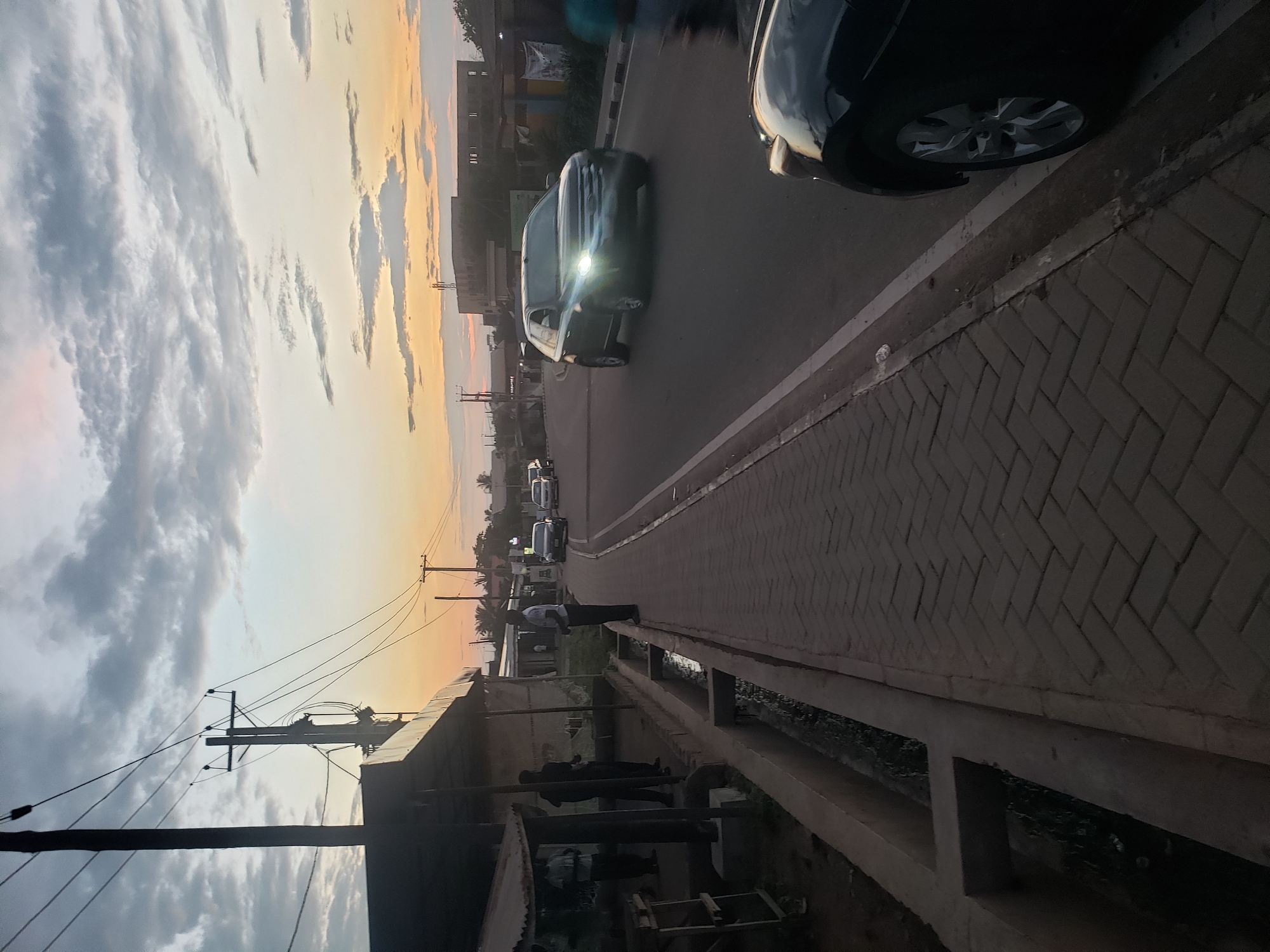 The image depicts a street scene at dusk in Narayi, Nigeria. On the left side of the street, there is a covered market stall with several people standing beneath it. A drainage channel runs alongside the sidewalk. The sidewalk is paved with interlocking bricks. The street itself is paved and appears to be a two-lane road. Several vehicles are present, including a dark-colored SUV with its headlights on driving down the street, and the front of a black car is visible in the lower right corner of the frame. In the background, there are buildings, utility poles with overhead wires, and trees. The sky is filled with clouds and shows the soft colors of sunset, transitioning from orange and yellow near the horizon to blue and gray higher up.
