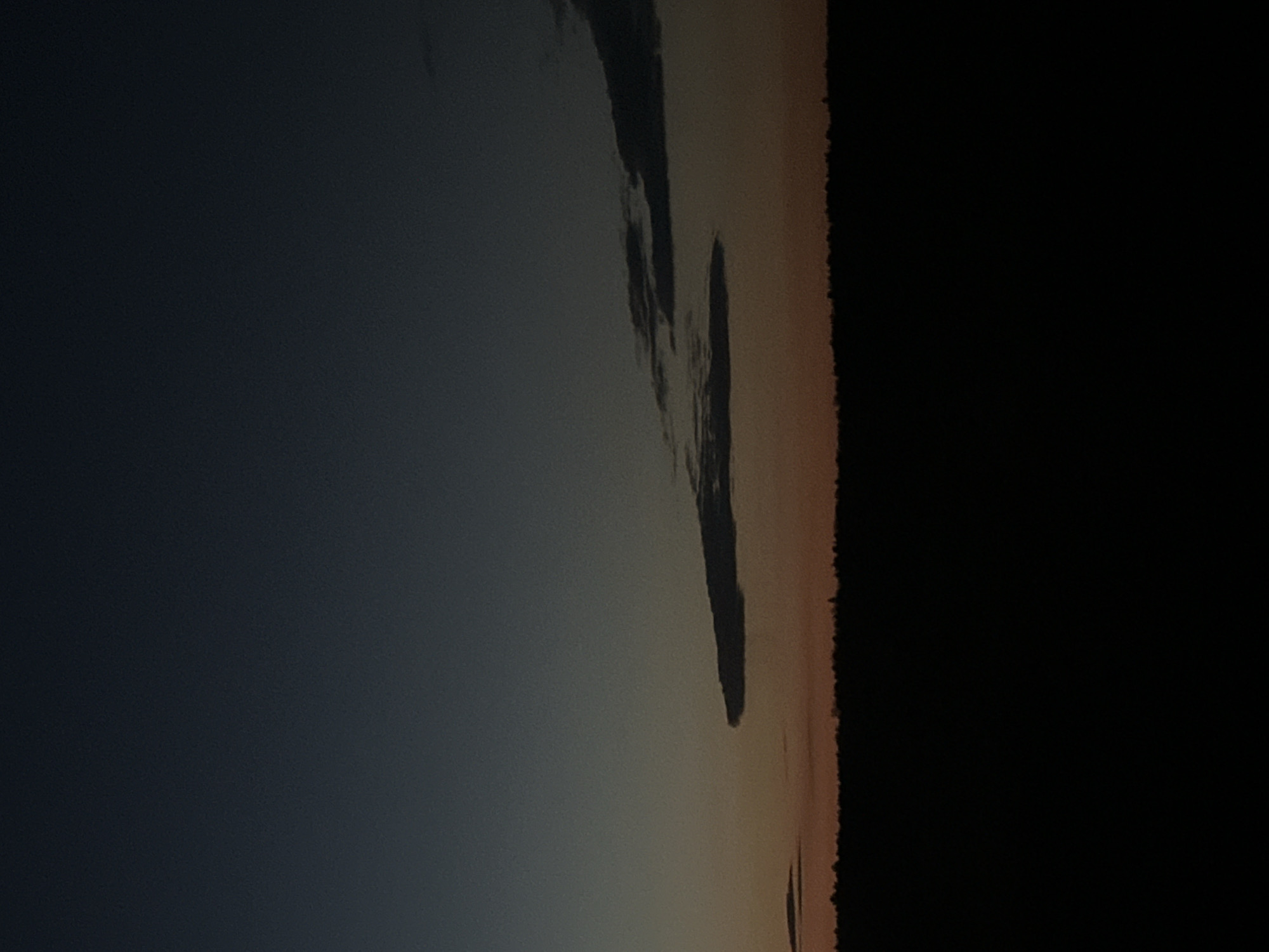 This landscape image, captured in Kharnavy, Russia, presents a serene twilight scene, likely at dawn or dusk. The horizon stretches horizontally across the lower third of the frame, revealing a dark, silhouetted landmass, which appears to be a distant forest or shoreline, against the ambient light. The sky above displays a striking gradient, transitioning from a deep, somber blue-grey in the upper left to a warm, soft orange-brown hue near the horizon line, indicative of the sun's low position. Prominent elongated, dark cloud formations are visible in the middle ground, appearing as distinct, shadowy streaks against the lighter parts of the sky, adding visual depth. No people or man-made objects are discernible, emphasizing the natural elements of the vast landscape. The atmosphere is tranquil and evokes a sense of natural beauty. The weather appears clear with these notable cloud structures. There is no visible text in the image.