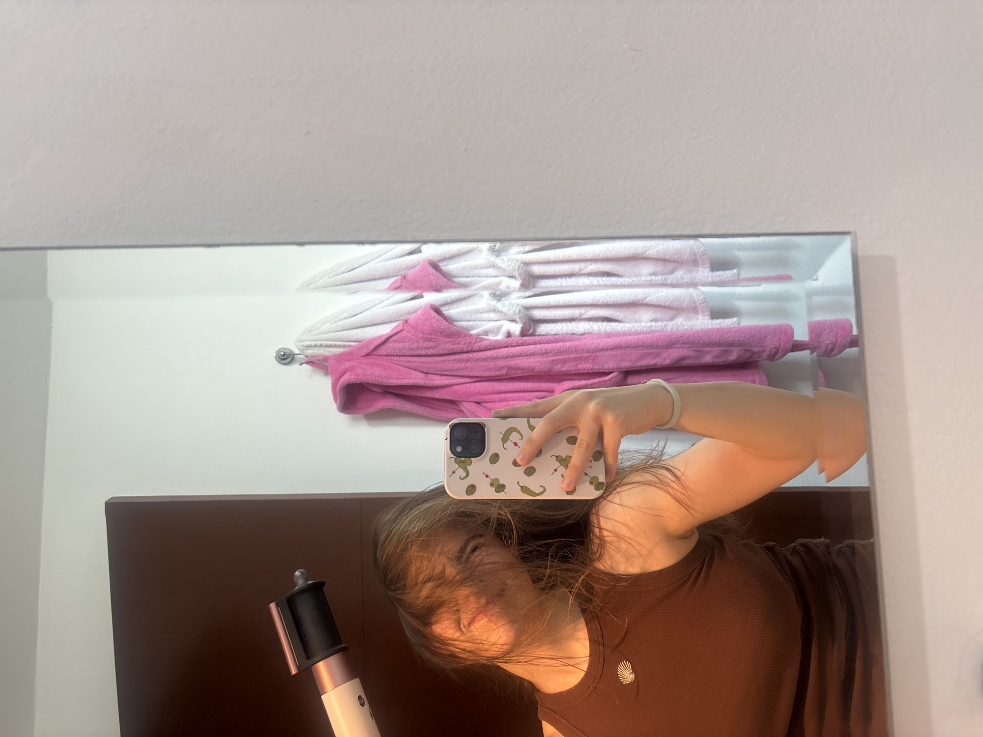 A mirror selfie captures a young person, likely in Puebla, Mexico, looking downwards with their light brown hair partially obscuring their face. They are wearing a dark brown, ribbed tank top or t-shirt, adorned with a light-colored, shell-shaped pendant on a necklace. A white bracelet is visible on their left wrist.
The person is holding a white iPhone, likely a Pro Max model due to its distinctive triple camera array, which is encased in a cream-colored phone case decorated with a repeating pattern of green chilies/peppers and green olives. The phone is held up towards the mirror, capturing the reflection.
In the lower-left corner of the image, a white and rose gold (or copper) hair styling tool, resembling a Dyson Airwrap, is partially visible.
The background, seen in the mirror's reflection, reveals an indoor setting, consistent with a bathroom or bedroom. Above the person's reflection, neatly folded white and pink towels are stacked on a white shelf or rack. A dark brown wooden headboard or wall panel is visible behind the person in the reflection, and a light-colored wall surrounds the mirror. The overall impression is a casual, domestic scene. No decipherable text is visible on any objects in the image.