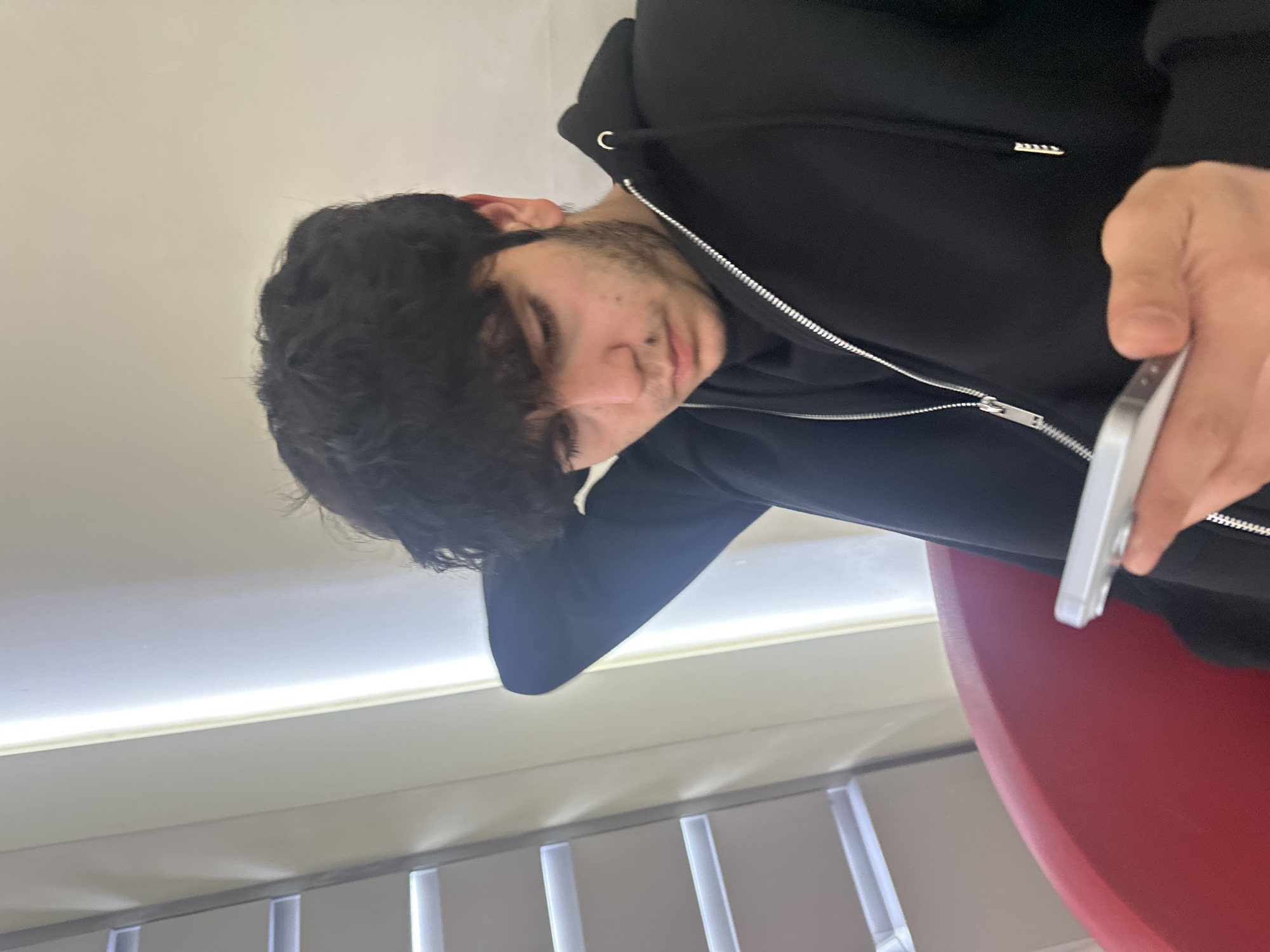A person with dark hair and a beard is looking at a cell phone. The person is wearing a black hoodie with a silver zipper. The background appears to be a room with white walls, a window, and a red curved object. The location and city are unknown.