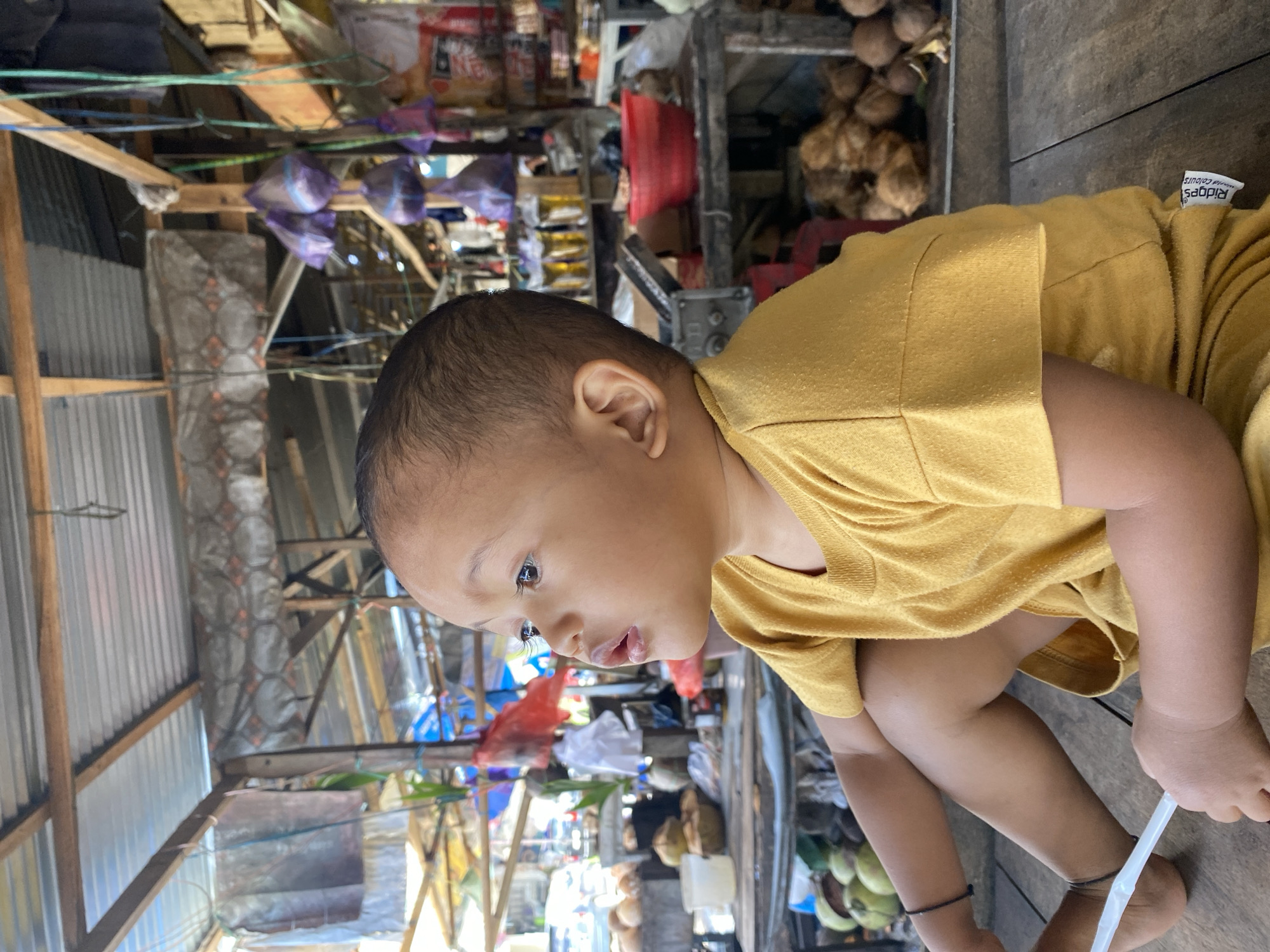 A young child in a yellow t-shirt sits on a wooden surface, looking to the left of the frame. The child's legs are crossed, and they are holding a white object in their right hand. The background shows a market scene with stalls made of wood and corrugated metal. Various items are visible, including coconuts, bags of produce, and what appear to be bottles. The overall impression is of a bustling outdoor market. The location is identified as Ta', Indonesia.