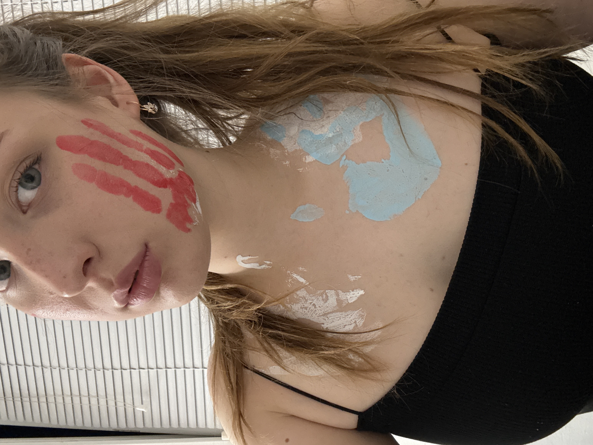 A young woman, with fair skin and light brown hair, has red paint applied in the shape of a handprint on her right cheek. She is wearing a black tank top. There are smudges of white and light blue paint on her chest and collarbone. Her left eye is visible, as is her nose and mouth. She has a small gold earring in her right ear. The background appears to be a window with horizontal blinds. The image is a close-up shot, focusing on the woman's upper body. The location in the city of Chaikovsky, Russia, is not depicted or suggested by the image.