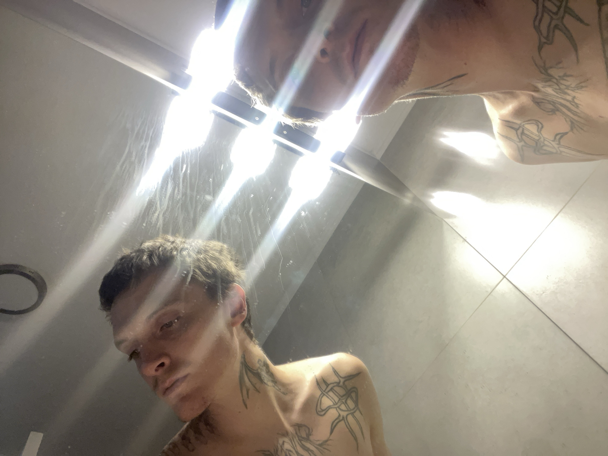 A shirtless man with light brown hair and several tattoos is visible in a mirror. He is positioned in what appears to be a shower or bathroom with white tiled walls. Overhead lighting creates a bright reflection in the mirror, obscuring the upper portion of the man’s reflection. The ceiling is also visible above the mirror's reflection. The location is not discernible from the image. Vicente López, Argentina is not identifiable from the visual information provided.