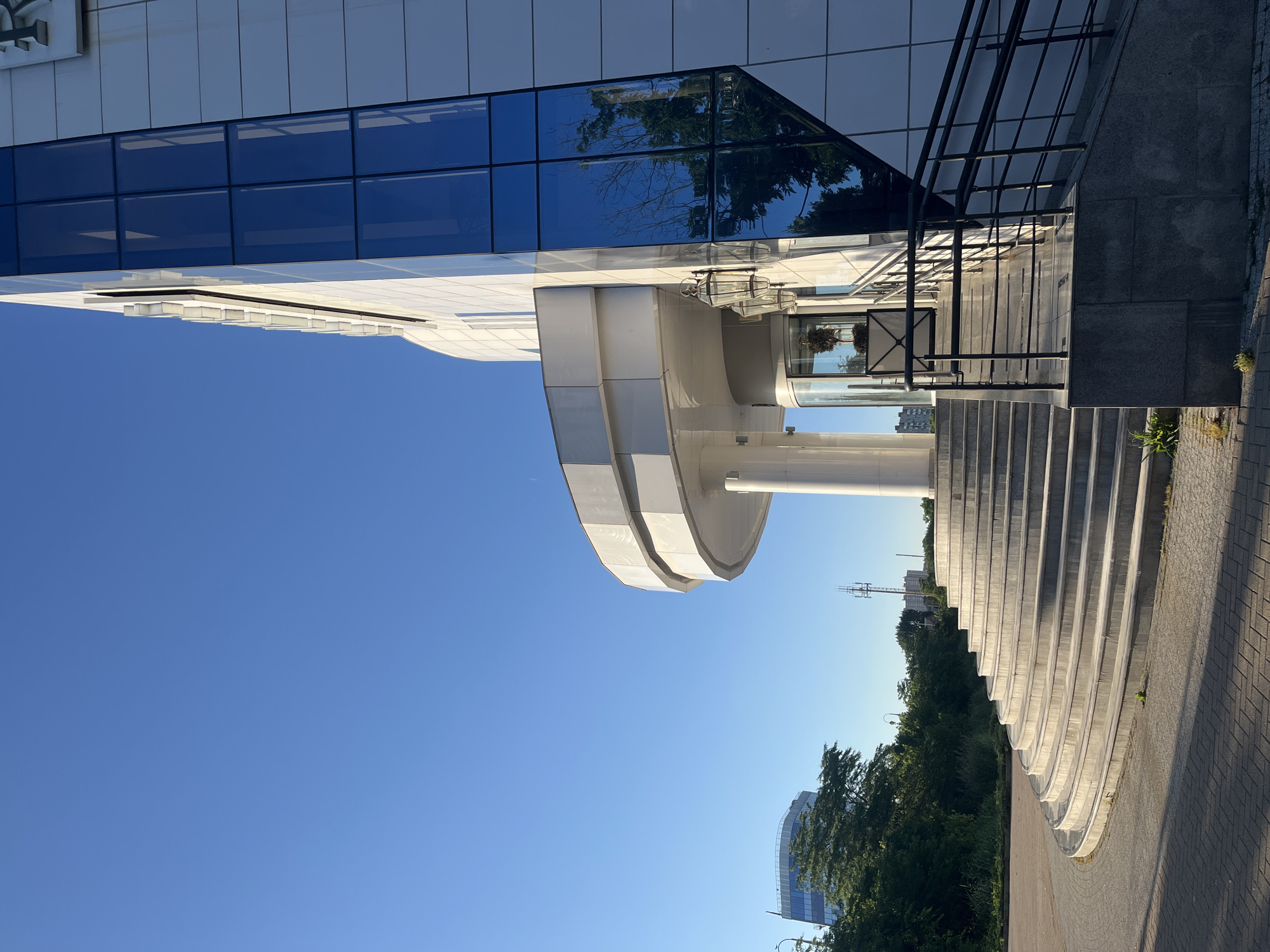 An exterior shot captures a modern, multi-story building with a white facade and a prominent glass section. A wide, curved staircase leads up to the building's entrance, which features a revolving door and decorative lanterns. The scene is set against a clear blue sky, with trees and other buildings visible in the background. The image appears to be taken in the city of Krasnodar, Russia, as suggested by the presence of specific architectural styles and urban elements characteristic of the region.