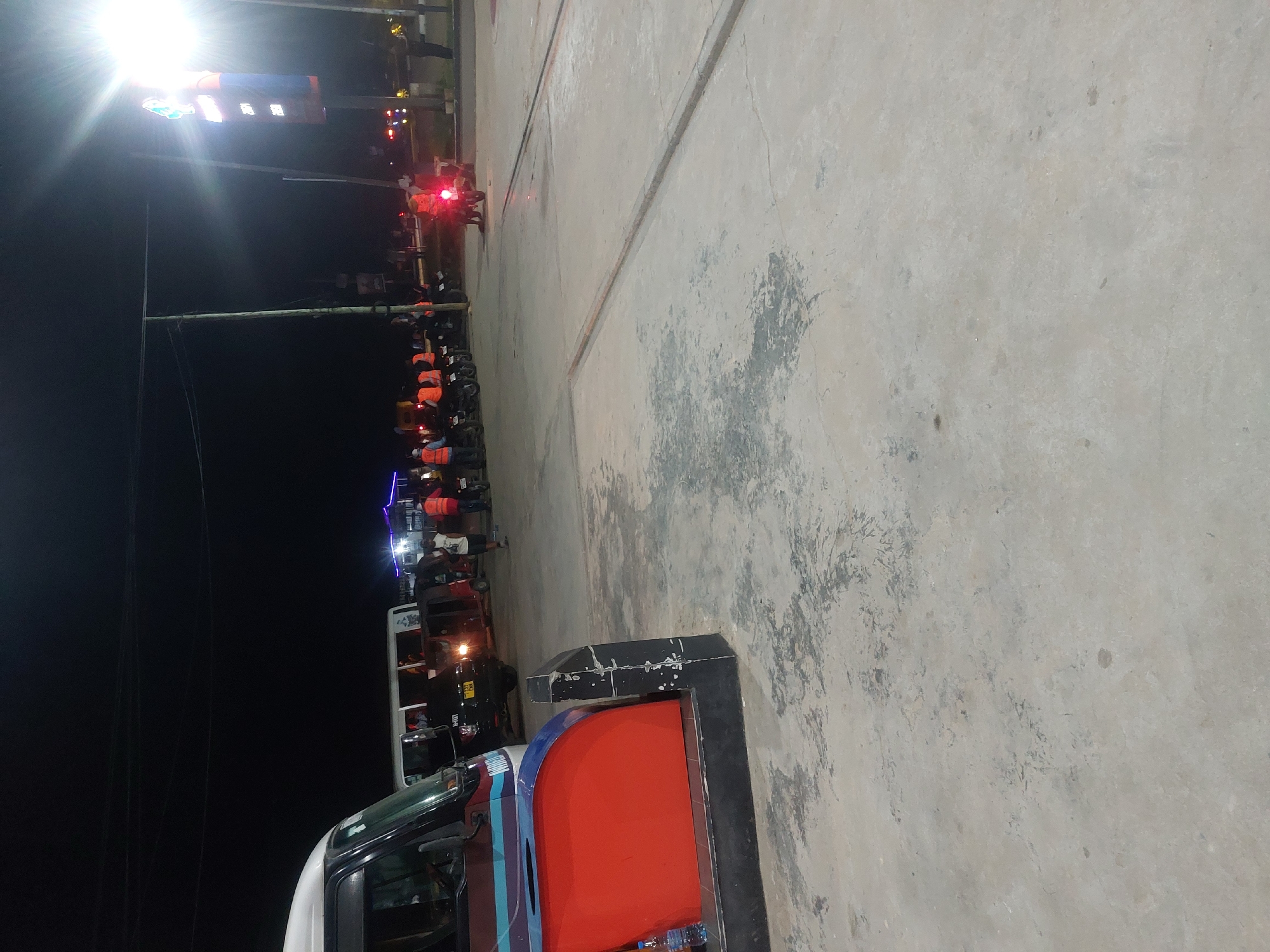 The image depicts a scene at night in Dar es-Salaam, Tanzania. A line of "bodaboda" drivers (motorcycle taxis) wearing orange vests are gathered near a traffic light showing red. The drivers are waiting along the edge of a paved area, with the street and other vehicles visible. There's an illuminated sign, possibly a business advertisement. A vehicle with a red and blue stripe is parked in the foreground. The background is dark, with streetlights and some building lights visible.