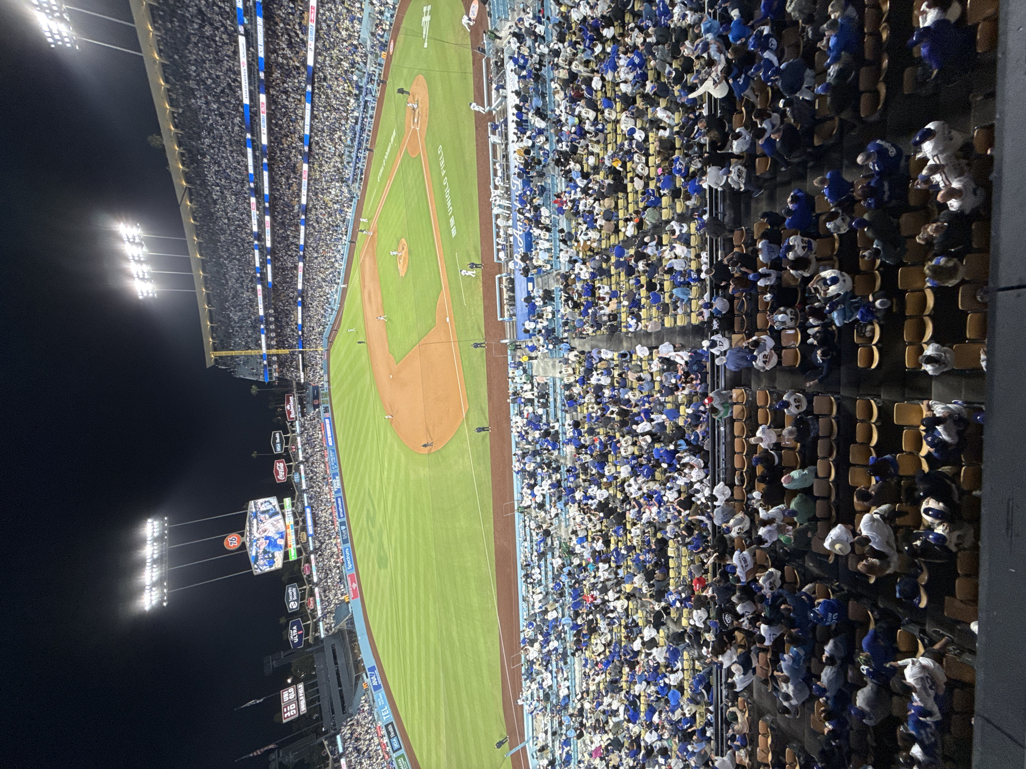 Here is a detailed description of the image:
The image depicts a night baseball game at Dodger Stadium, in Los Angeles, California, United States of America. The stadium is filled with spectators, the majority of whom are wearing Dodger blue. The baseball field is green and the infield is brown. Bright stadium lights illuminate the scene. Numerous advertisements are displayed along the stadium walls and on the scoreboard. The stadium is densely packed with fans, suggesting a well-attended game.