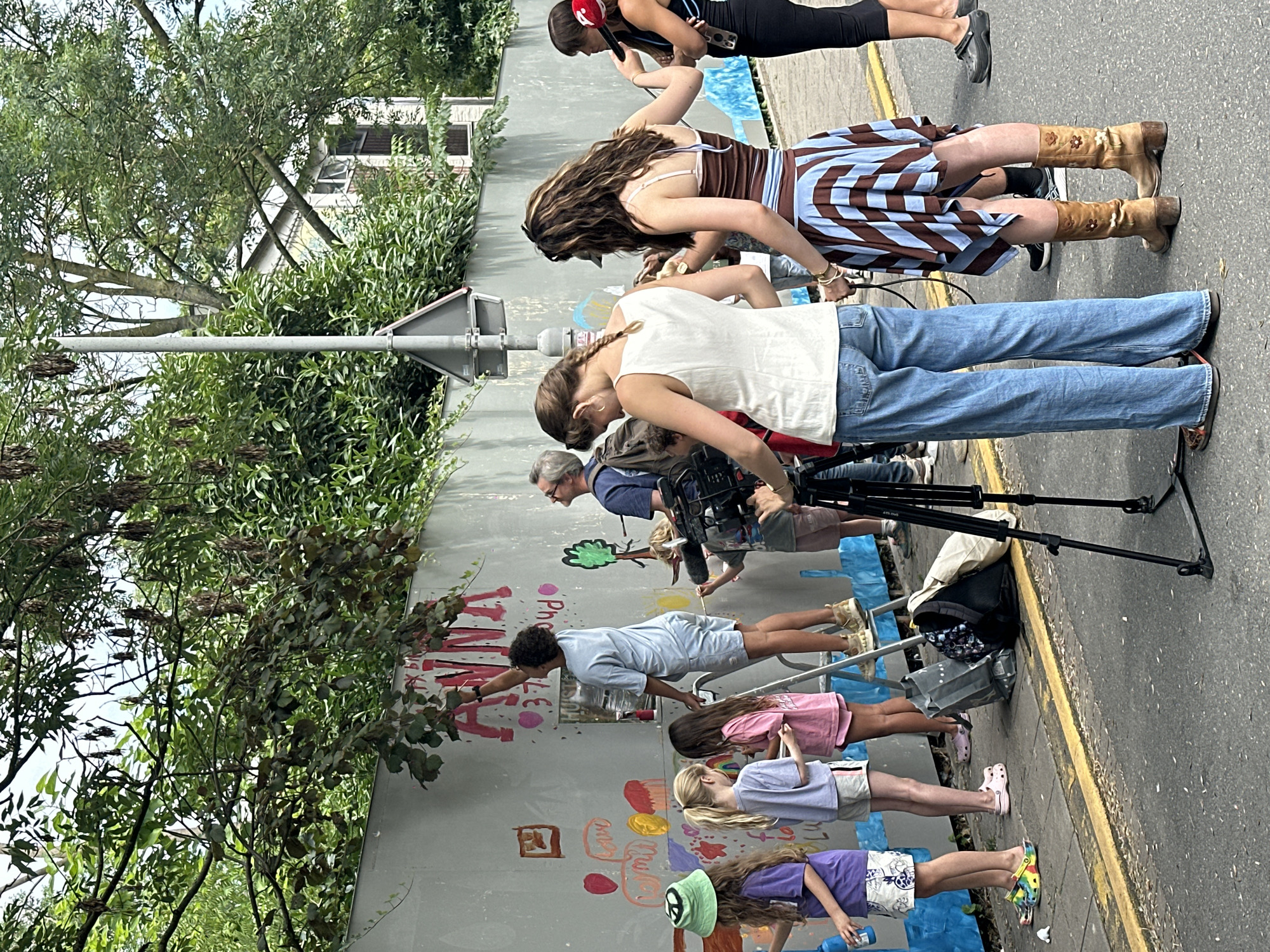 The image shows a group of people, including children and adults, painting on a concrete wall on a city street. The wall has various drawings and text, including the word "ANNA" in large pink letters. Some people are using paintbrushes and buckets, while others are observing or filming. A camera on a tripod is positioned in the foreground, suggesting a filming or documentation event. The environment appears to be an urban street with a sidewalk, a road, and trees and buildings in the background. The specific location in Amsterdam, Netherlands, is not explicitly indicated in the image.