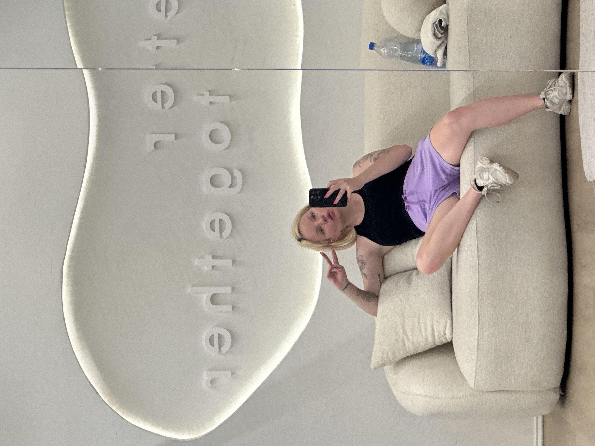 A person with blonde hair and tattoos on their arms is holding a phone and making a peace sign in a selfie. The person is wearing a black tank top and purple shorts and is sitting on a beige couch. The couch also has a water bottle and white towel. Behind the person is a large, white wall decoration with the word "together" embossed on it in a vertical stack. The image appears to be a reflection in a mirror, creating a double image effect. The mirror seems to be located in a store, potentially in Budva, Montenegro.