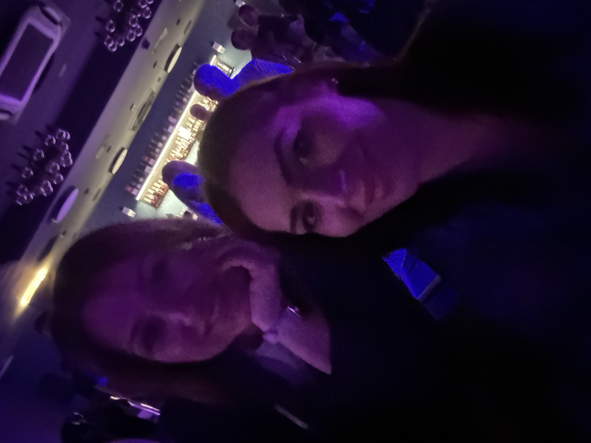 Two women are in a dimly lit bar in Kazanlak, Bulgaria. The background shows a bar area with bottles and hanging lights. Other people are visible in the background. The lighting gives off a blue/purple hue.