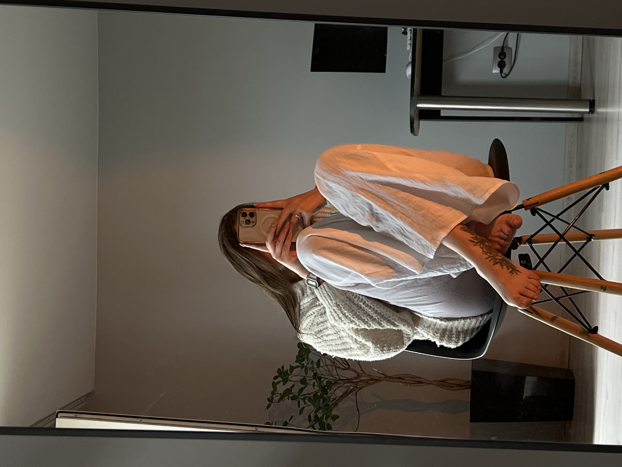 A person is seen in a mirror selfie, captured in what appears to be an indoor setting, possibly a room in a home. The individual, with long brown hair, is seated on a chair with a black plastic seat and light wooden legs with a black wireframe base. They are wearing loose-fitting white clothing, possibly a top or dress, and a cream-colored chunky knit cardigan or sweater. Their face is obscured by a light-colored smartphone they are holding to take the picture, revealing a simple band ring on one finger. A tattoo with a botanical or floral design is visible on their right foot/ankle, which is resting on a part of the chair's leg structure. A watch with a light-colored band is visible on their left wrist.
In the background, visible through the mirror's reflection, there is a gray wall with a corner junction. To the right, there's a dark-colored desk or table with a silver support leg or bar, and a wall-mounted power outlet with two round holes and a grounding pin is visible above it, along with some cables. A dark object, possibly a monitor, is also seen on the desk. A green leafy plant is partially reflected in the lower left portion of the mirror. The overall ambiance suggests a casual, relaxed moment. There is no visible text in the image.