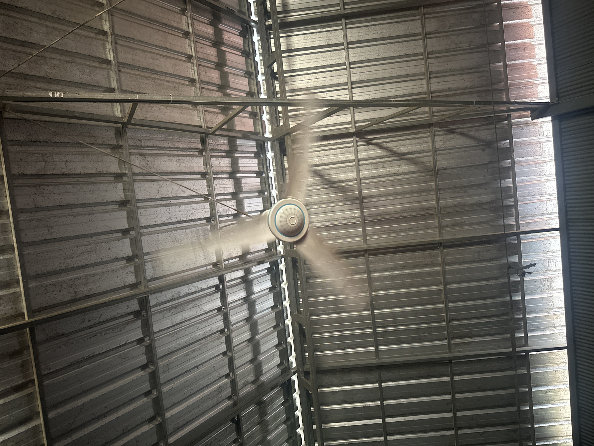 The image shows a ceiling fan in motion, mounted on a corrugated metal roof structure in what appears to be an industrial or warehouse-like setting. The fan blades are blurred due to their speed. The surrounding environment consists of metal beams, corrugated metal walls, and a corrugated metal roof. Sunlight is filtering in from the sides, creating bright areas and shadows. The scene is devoid of people. The location is identified as Thbong, Cambodia.