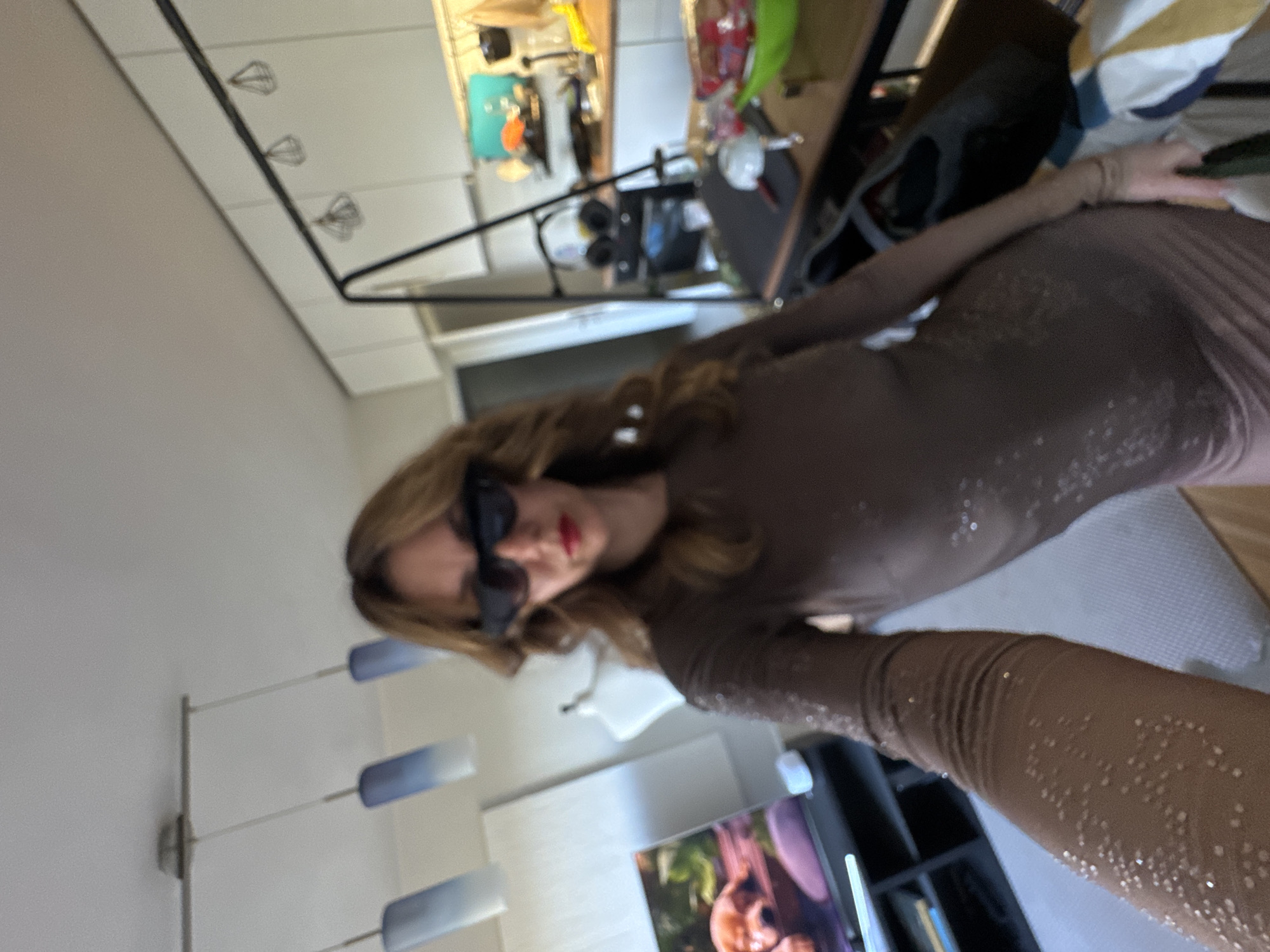 A woman wearing sunglasses and a sparkly brown dress is indoors. Behind her is a television, a bookcase, and a modern light fixture with blue shades. The background includes an out-of-focus kitchen area with cabinets and appliances. The specific location within Sofia, Bulgaria, is not discernible from the image.