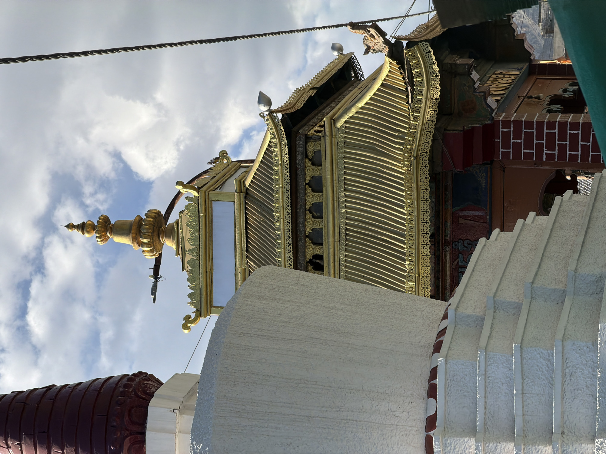 The photograph, taken in Leh, India, depicts a portion of the Leh Palace and its surrounding structures under a cloudy sky. In the foreground, a white stupa with tiered steps is visible, its textured surface illuminated by the sun. To the right and slightly behind the stupa, the ornate golden roof of a Tibetan-style building, likely part of the Leh Palace complex, dominates the midground. This building features multiple tiers of steeply sloped, golden roofs adorned with intricate carvings and decorative finials, including lotus-shaped elements and animal motifs. The facade of this building is a deep red and brown, with visible brickwork and decorative paintings. A dark, coiled cable runs vertically on the far right of the image, suggesting the presence of infrastructure. The background is filled with a sky characterized by scattered clouds, with a hint of mountains visible in the far distance. There are no people or visible actions in the scene.
