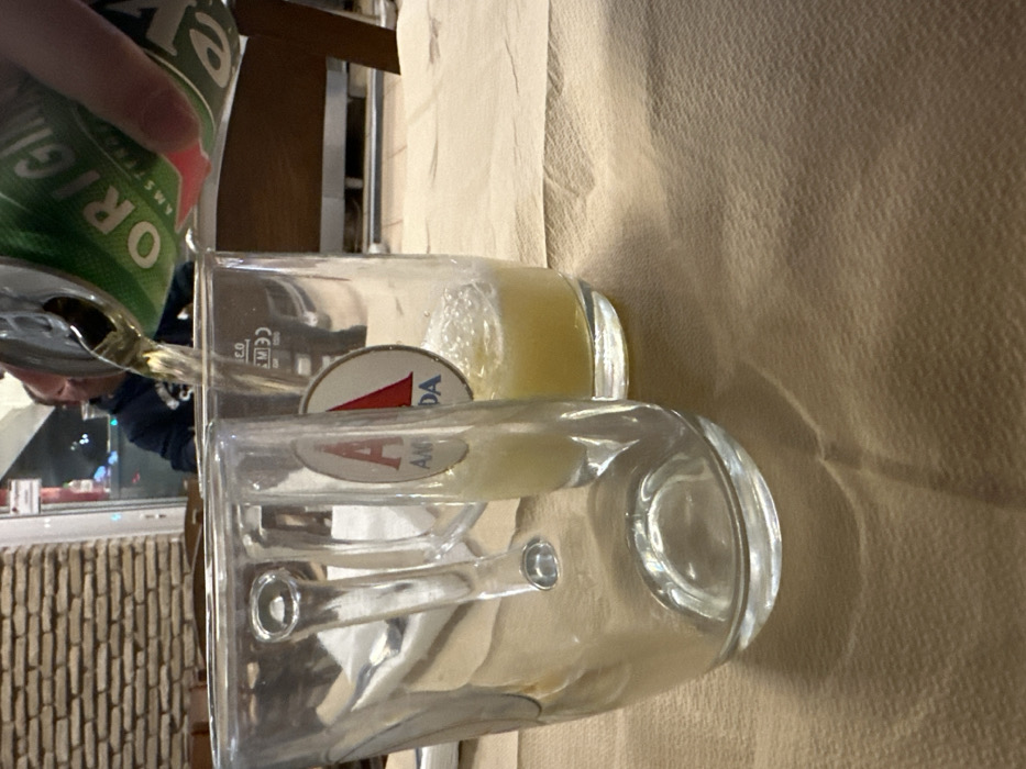 The image, taken in Nea Erithrea, Greece, is oriented sideways, depicting the act of pouring a beverage indoors. In the upper left, a person's hand holds a green and silver can, from which a golden liquid is actively being poured into a clear glass mug. The can has visible text including "ORIGINALS" and "AMSTERDAM," consistent with a Heineken brand.
Two clear glass mugs are central to the composition. The mug receiving the pour, along with a second mug positioned behind it, both feature a distinctive red and white logo with a prominent "A," followed by "DA," and "AMSTERDAM" beneath, identifying them as Amstel branded glassware. The mug currently being filled also has "CE M03" and "0.3 L" etched into its side, indicating its capacity. The second mug contains a small amount of the golden liquid already settled at the bottom.
The mugs rest on a light brown, subtly textured surface, likely a napkin or tablecloth. Reflections on the glass surfaces show elements of the surrounding indoor environment, including what appears to be a person's reflection in the background and a textured wall. The lighting suggests an evening or nighttime setting.