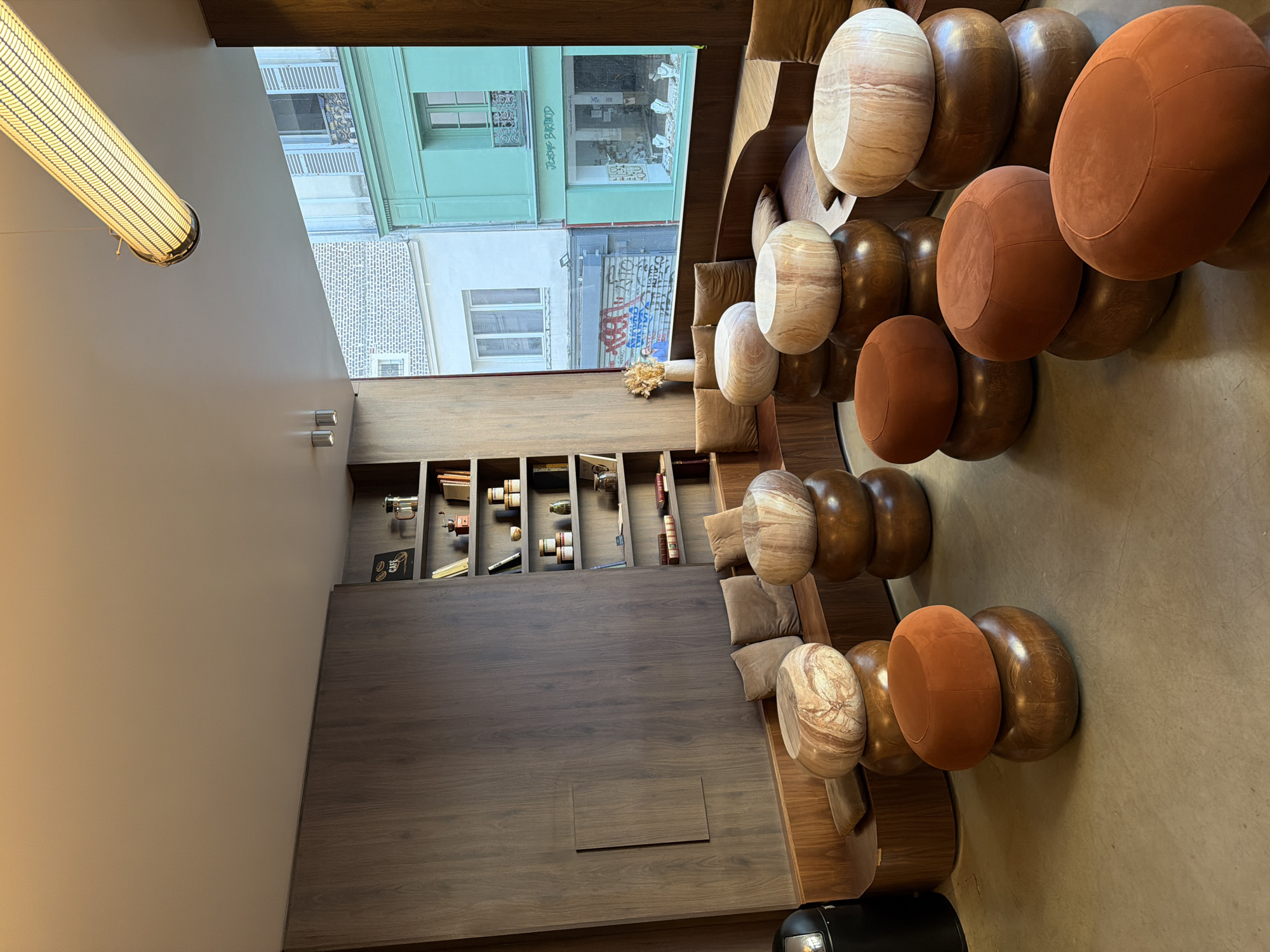 Captured from an elevated, slightly tilted perspective, the image presents the interior of a cafe or restaurant in Paris, France. The interior features a modern design, with a row of rounded wooden and upholstered stools and chairs aligning the right side of the frame. A long, built-in wooden bench with cushions extends along the lower portion of the frame, leading towards a wall-mounted bookshelf containing various decorative objects and books.
Large windows offer a view of the Parisian cityscape, revealing neighboring buildings with architectural details and signage that appears to read "JEROME BILLAUD." The external scene also includes graffiti-covered walls.
A cylindrical light fixture hangs from the ceiling on the left side of the image, illuminating the interior space. The color palette is primarily composed of warm browns and neutral tones, contributing to a cozy atmosphere. There are no people depicted in this image.