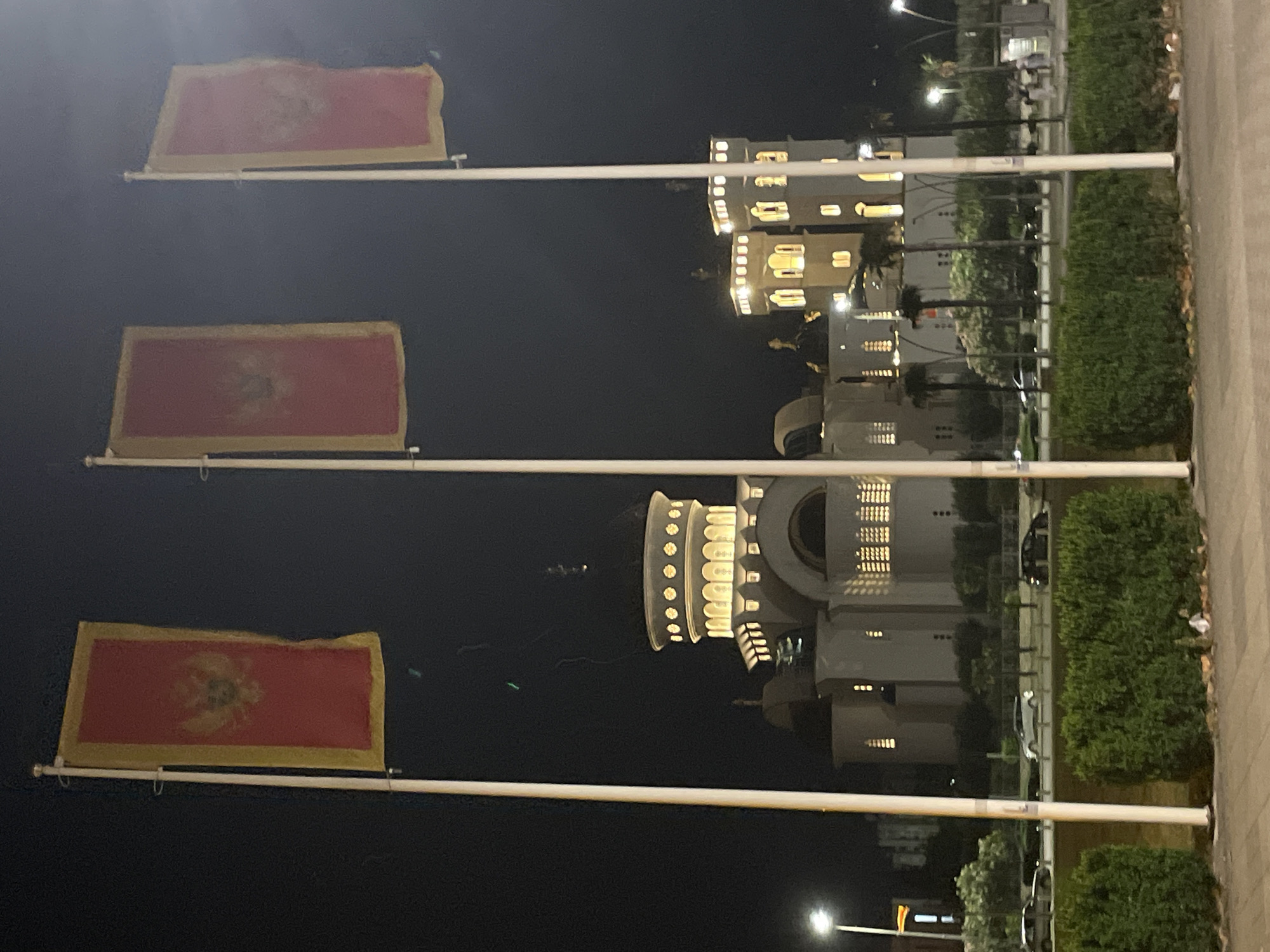 At night, three Montenegrin flags fly in front of a light-colored building with lit windows in Bar, Montenegro. The flags are red with a gold border and a gold emblem. The building is tall and has a rounded section with many small lights. A row of trees separates the building from a sidewalk. Street lights are visible in the background.