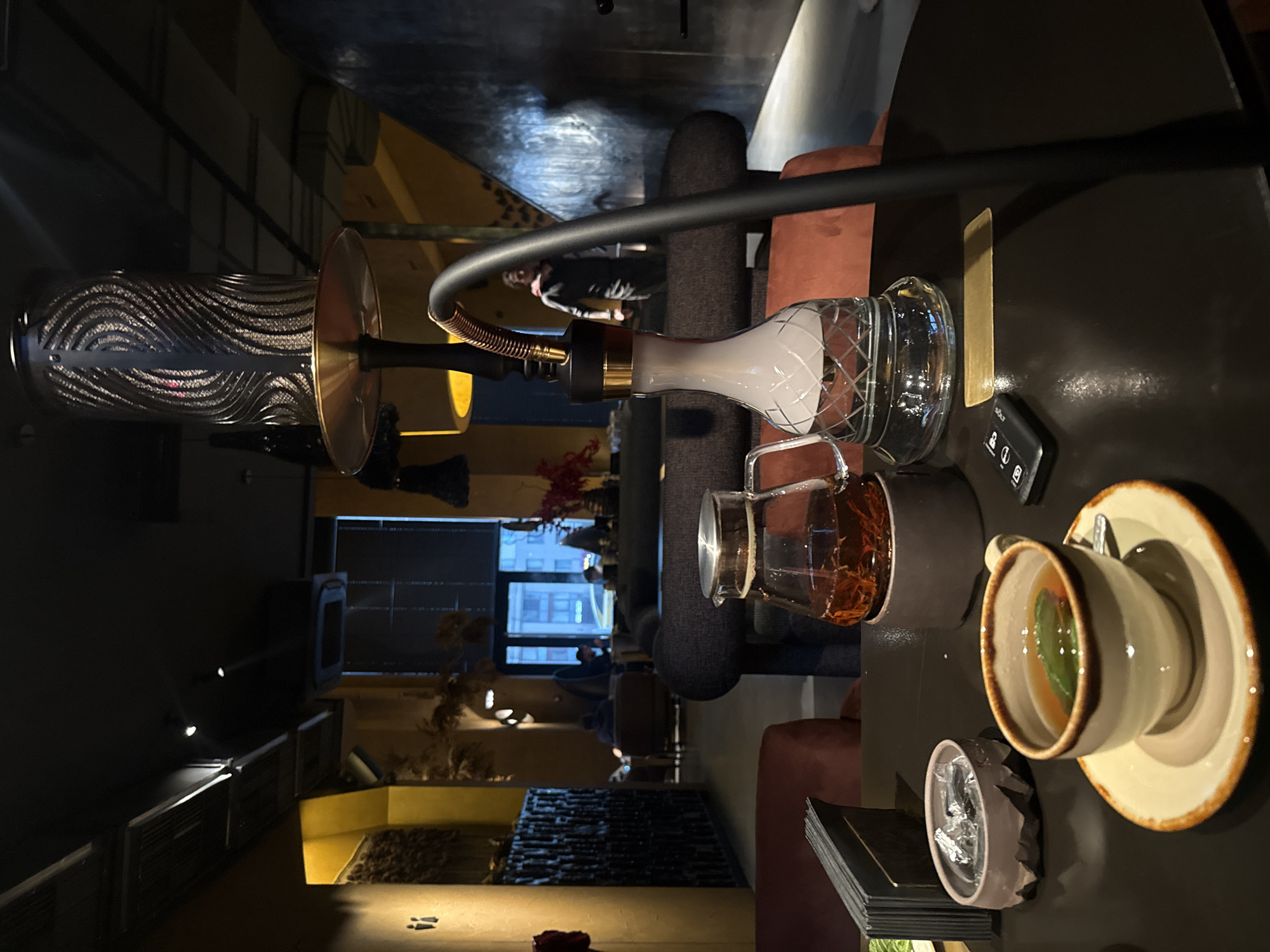 A high-angle shot depicts a table setup in what appears to be a dimly lit lounge or bar, likely within the city of Khimki, Russia, given the prompt context. A hookah, including a patterned hose, a water base with ice, and a central stem, takes a prominent position on the table. The table also hosts a cup of tea with a saucer, a glass of amber-colored liquid, a small container with what seems to be ice, and what may be a set of menus or cards. In the background, seating arrangements and indistinct figures suggest a populated space, possibly a restaurant or lounge. Natural light filters in through a window in the backdrop. The scene seems to capture a relaxed, social setting.