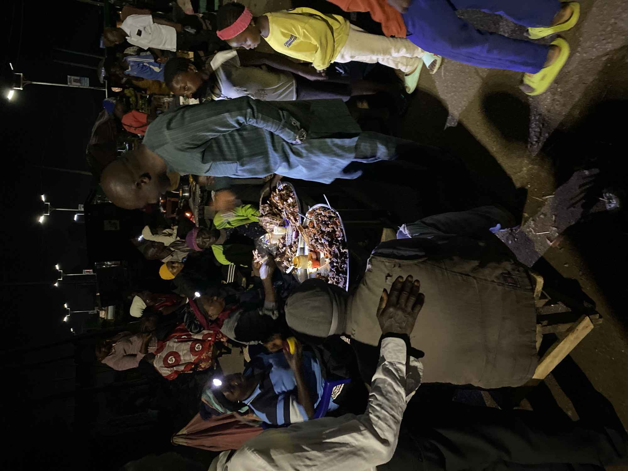 A nighttime scene under streetlights shows a group of people gathered around a table laden with unidentified food items, potentially in Bukuru, Nigeria. Individuals wear headlamps, suggesting limited lighting. People stand and sit, eating. The scene has a sense of community with visible interactions.