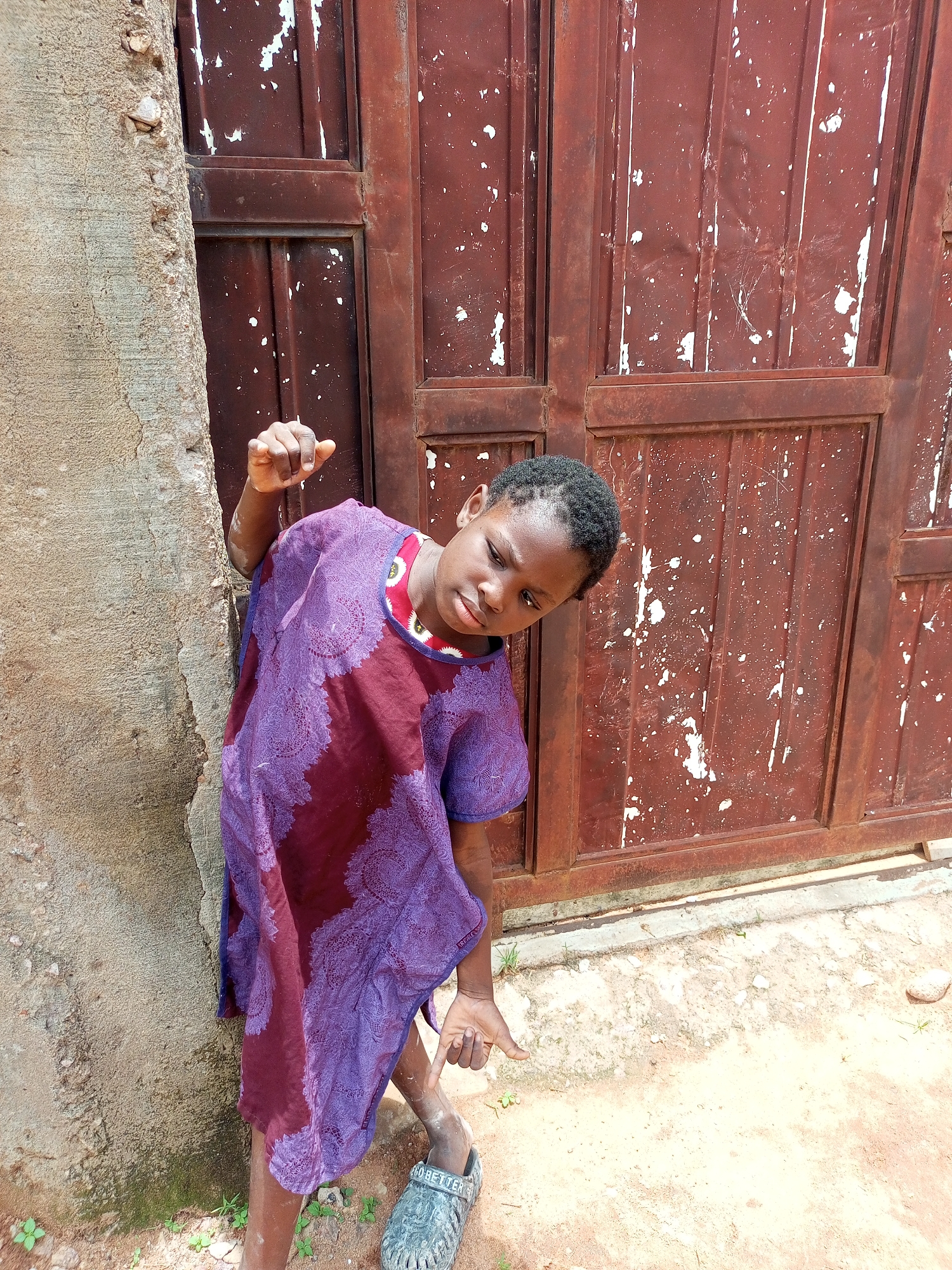 In Tempure, Nigeria, a young girl with braided hair, wearing a purple and maroon patterned garment, leans against a rough, textured concrete wall. She is positioned to the left of a weathered, maroon metal door, which shows signs of age with peeling white paint. The ground in front of the door is a mix of dirt and sandy soil, with sparse green weeds and a few small stones. The girl is looking down and to her right, with her left arm bent upwards and her fist clenched, while her right hand is extended downwards, pointing towards the ground. She is wearing a dark-colored sandal on her visible left foot. The overall setting appears to be an outdoor, possibly residential, area.