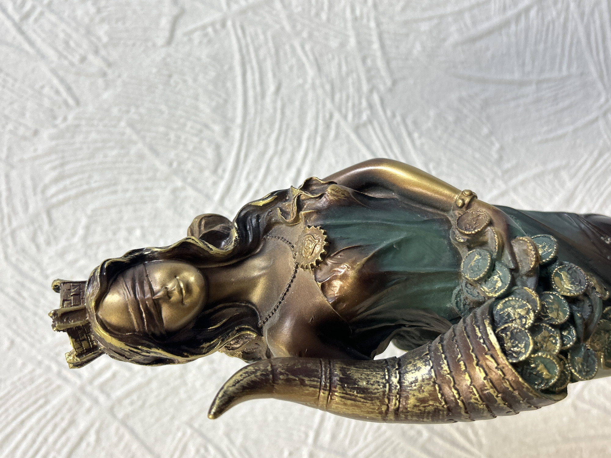 The image displays a close-up, horizontal view of a bronze or bronze-finished statue, set against a textured white background. The central subject is a female allegorical figure, possibly representing abundance, fortune, or Lady Luck.
The figure's eyes are blindfolded, suggesting impartiality or a focus on inner perception. She wears an elaborate, crown-like headpiece and has long, flowing hair. Her body is draped in flowing garments, with a significant portion colored in a dark green, providing a contrast to the metallic bronze finish of the rest of the statue. A delicate necklace with a distinctive, gear-shaped pendant adorns her chest.
A prominent feature of the statue is a large cornucopia, or horn of plenty, which appears to be held by or integrated with the figure. This cornucopia is depicted overflowing with numerous coins or medallions, each detailed with an unidentifiable design. These coins, like parts of the drapery, also incorporate shades of green and bronze.
The background is a simple, textured white surface, possibly fabric or a wall, which ensures the statue is the sole focus. There is no visible activity or event, as the image captures a static art object. No specific time of day, weather conditions, or discernible text are present within the frame. The statue's overall appearance conveys themes of prosperity and destiny. The provided location context is Chaikovsky, Russia, but this is not visually indicated in the image itself.