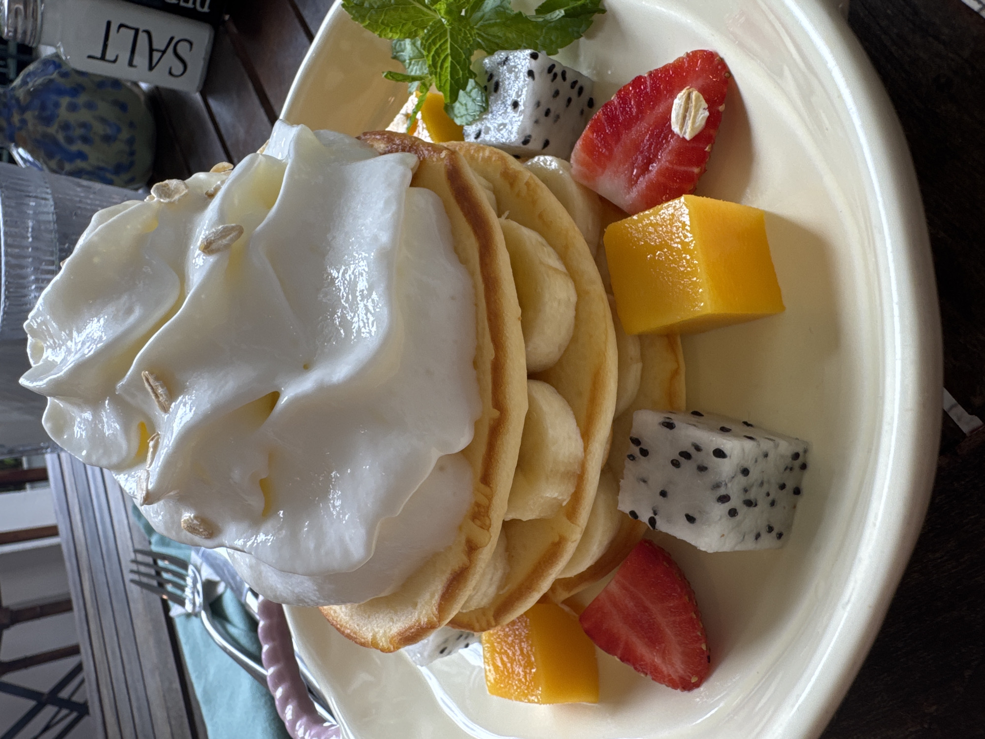 The image shows a plate of pancakes topped with a large dollop of whipped cream, arranged with slices of banana between the pancakes. Cubes of mango and dragon fruit, along with sliced strawberries, garnish the plate. A sprig of mint is also present. A fork and knife rest to the side of the plate on a light green napkin. A salt cellar is partially visible in the background. The context suggests the image was taken in Đà Nẵng, Vietnam.