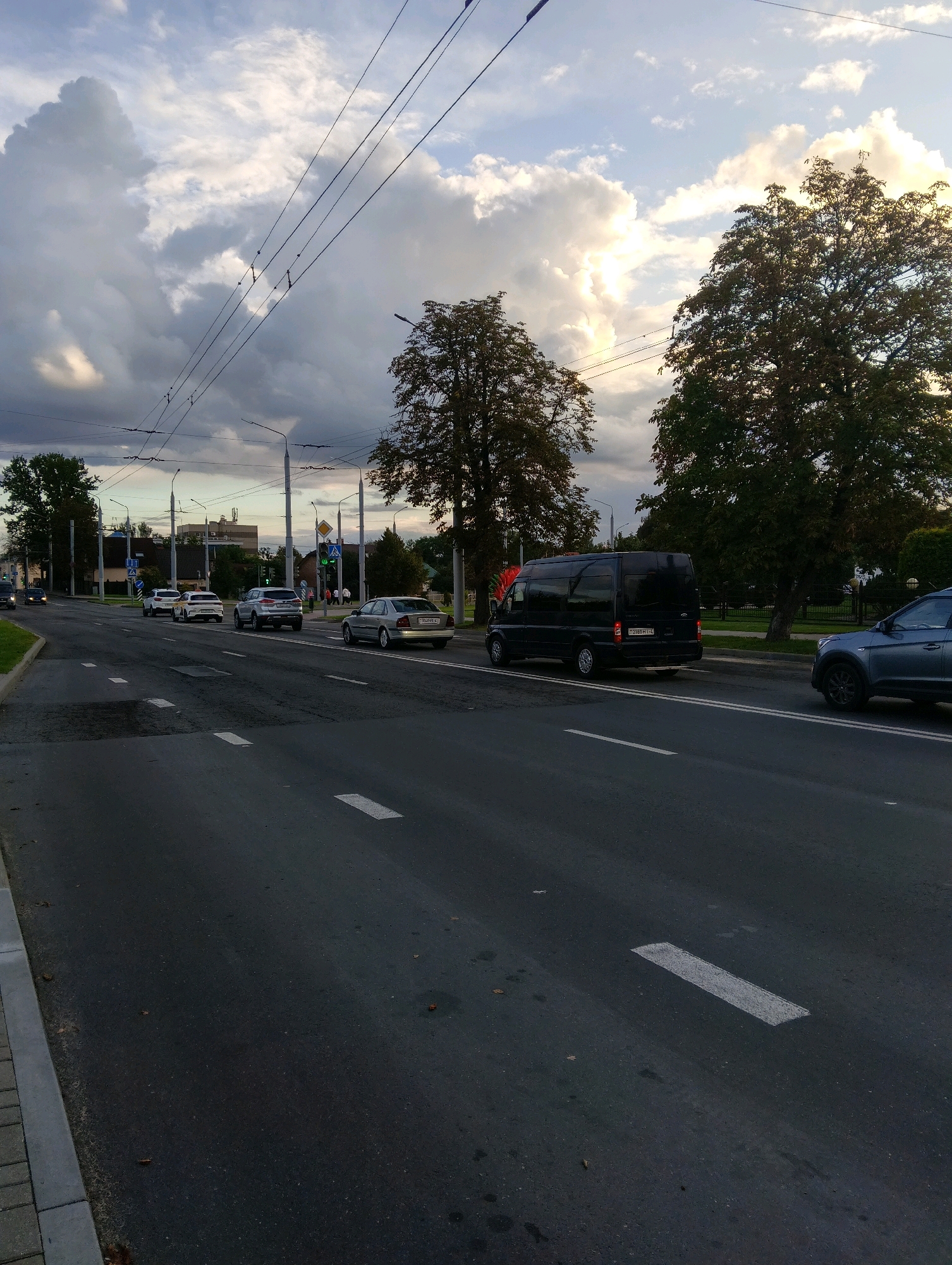 The image captures a street scene in Hrodna, Belarus, under a cloudy sky with patches of sunlight. The perspective is from the side of the road, looking down a multi-lane asphalt street. Several cars are visible, including a silver sedan, a silver SUV, and a black minivan. Overhead, power lines and streetlights are strung across the street. Trees line the sides of the road, and buildings are visible in the background. The street has white lane markings. The overall impression is of a typical urban environment during daylight hours.