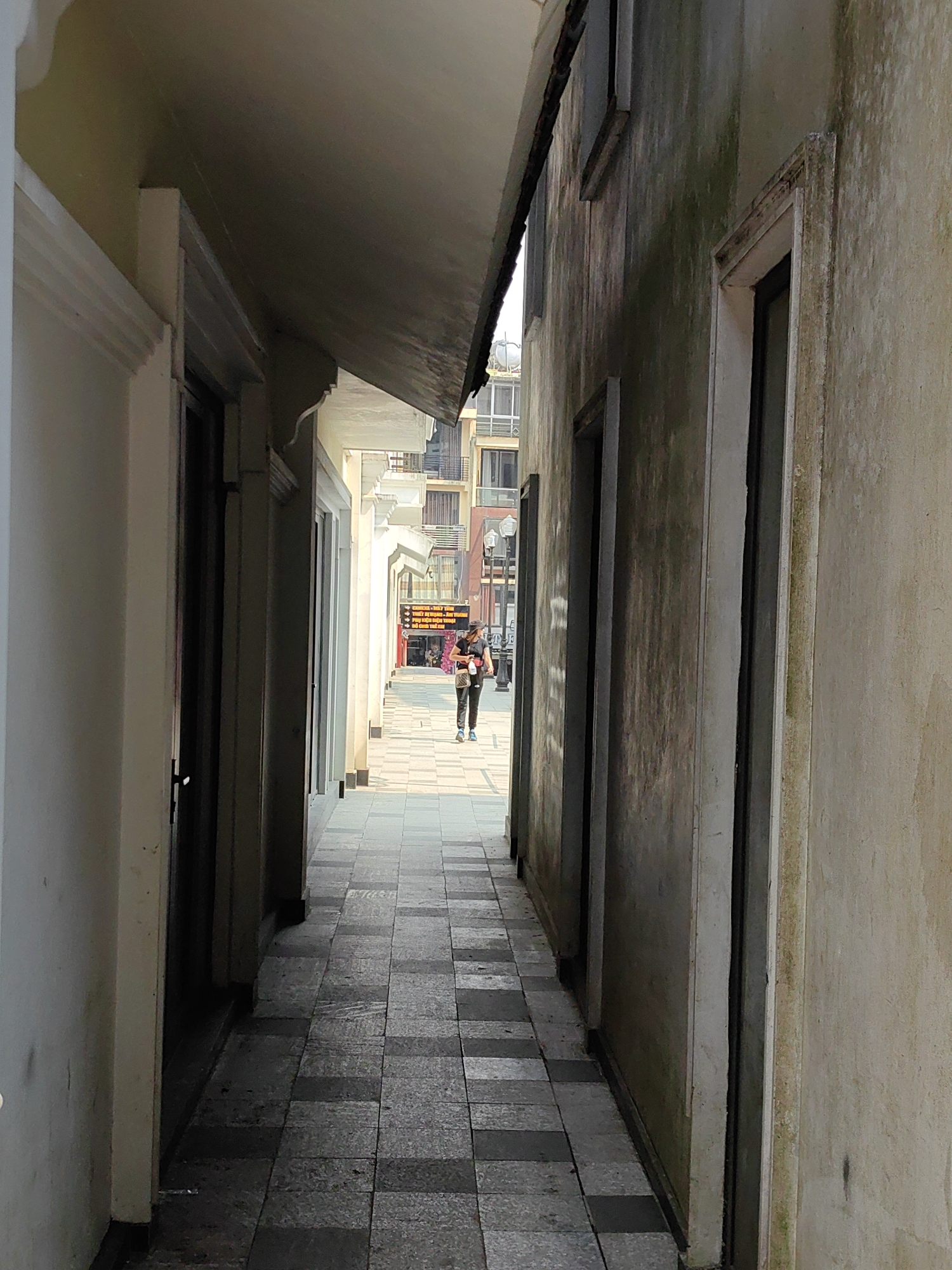 A narrow pedestrian passage in Sa Pa, Vietnam, is depicted, extending from a shadowed foreground towards a brightly lit street in the distance. The passage floor is paved with an alternating pattern of dark and light grey square tiles. The left wall is smooth, light beige, featuring multiple recessed doorways or windows and an overhead awning. The right wall is textured, light grey, showing extensive dark green and black biological growth, also containing recessed openings and an overhead awning.
At the far end, the passage opens onto a wider urban street lined with multi-story buildings, some featuring balconies. Two street light poles are visible on the left side of the street. Prominent commercial signage includes a large yellow sign with black Vietnamese text (e.g., "MÁY TÍNH," "ĐIỆN THOẠI," "TIVI") and a red sign positioned below it.
One individual is observed walking away from the camera, towards the illuminated street. This person wears dark trousers, a dark top, a dark wide-brimmed hat, and light blue athletic shoes. A light-colored bag is carried on their right side. No other individuals or interactions are present in the immediate scene.