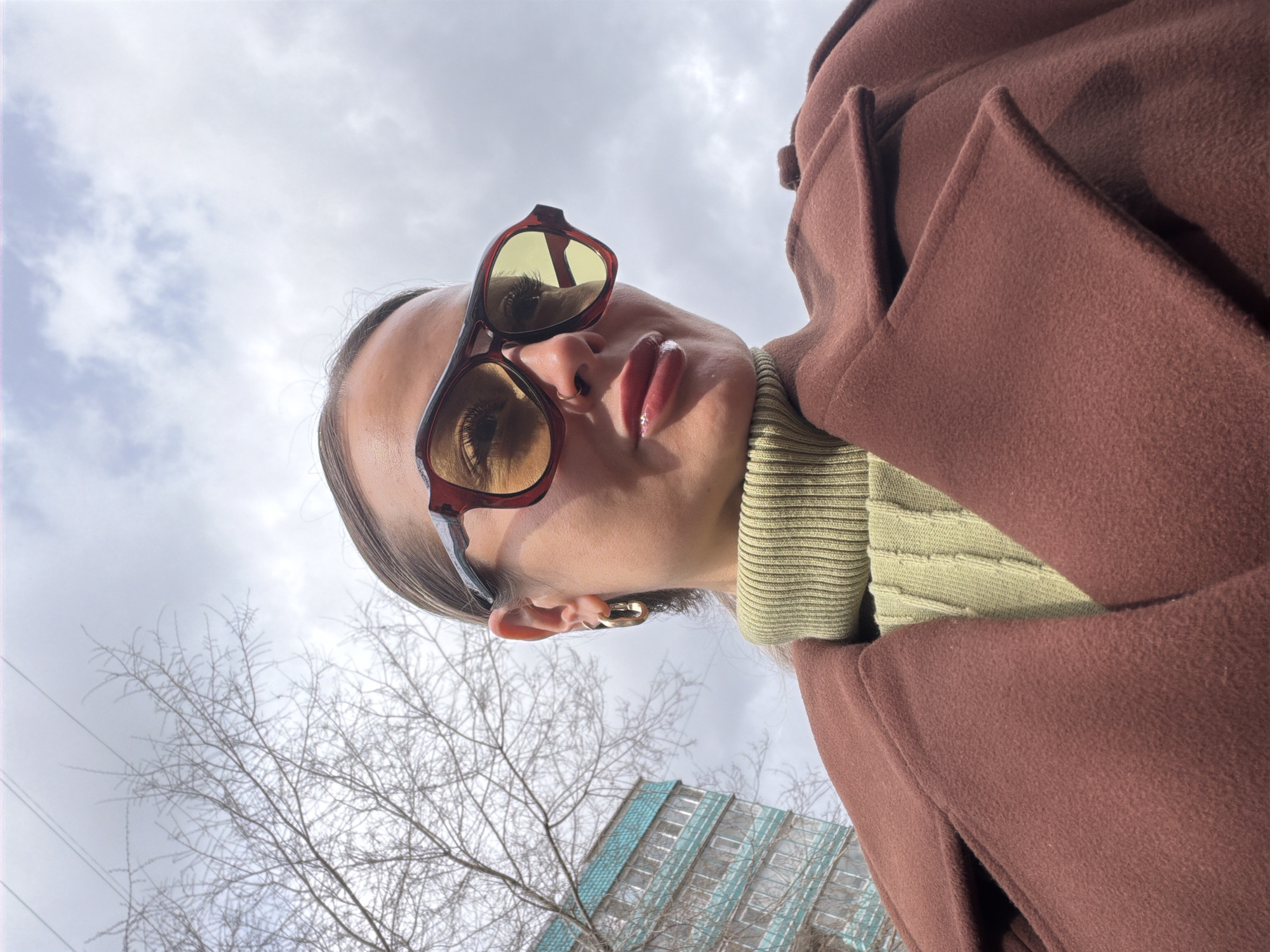 A woman wearing sunglasses, a green turtleneck sweater, and a brown coat is the main subject of the image. She is positioned in front of a building with blue and white horizontal stripes, suggesting an urban environment. Bare tree branches are visible against a cloudy sky. The photo is taken from a low angle, tilting upwards towards the woman. The building in the background may be located in the city of Krasnoyarsk, Russia.