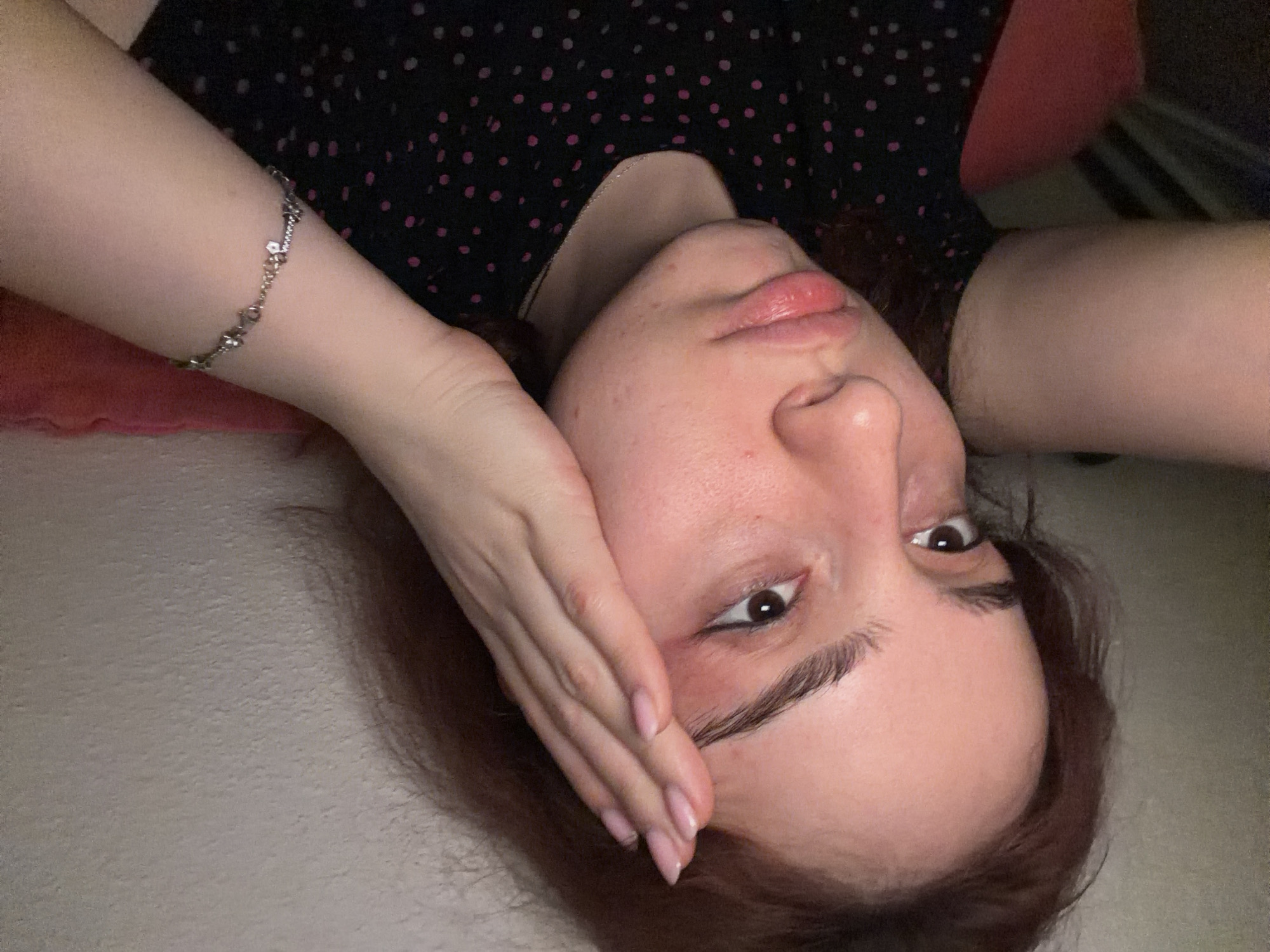 The image is a close-up, head-and-shoulders portrait of a young person with reddish-brown hair and fair skin. The person is looking towards the viewer with a pensive expression. Their right hand is raised to their forehead, with fingers splayed. A silver-toned bracelet with small charms is visible on their left wrist. They are wearing a black top with a pink polka dot pattern. The background is indistinct, appearing to be a textured wall and possibly a piece of furniture. There is no visible indication of the city of Strasbourg, France, or any specific urban environment.
