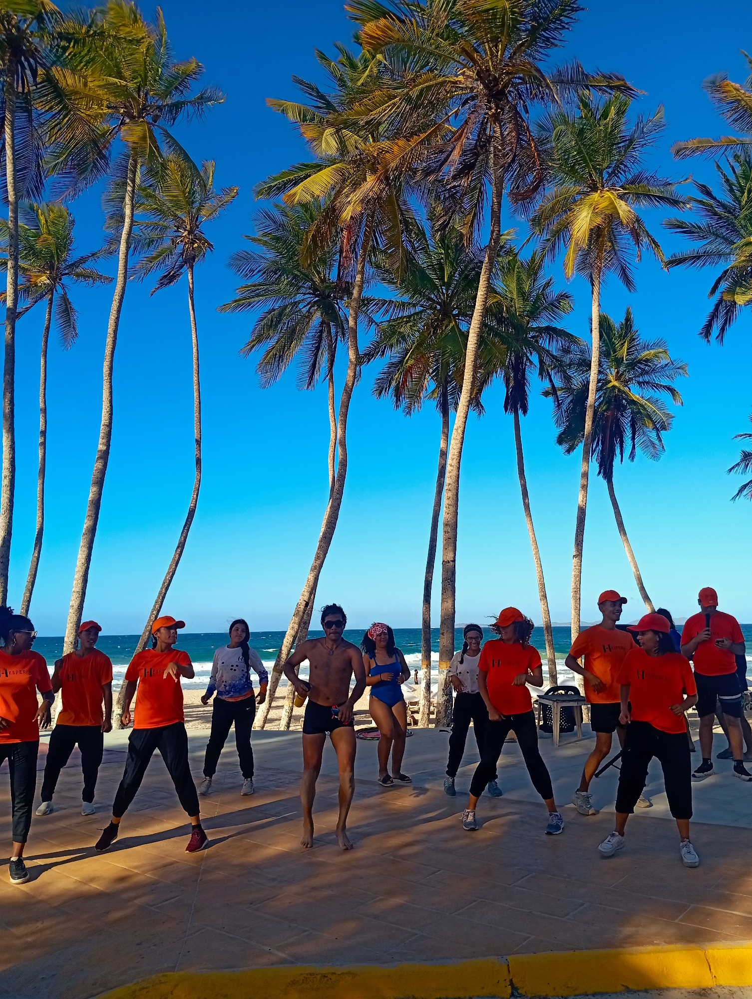 On a sunny day at Playa El Agua, Venezuela, a group of people are engaged in a fitness activity outdoors. Predominantly, ten individuals wearing orange T-shirts and matching caps are seen performing synchronized movements on a paved surface. Interspersed among them are three individuals in other attire, including a man in swimwear. The backdrop is composed of palm trees against a clear, bright blue sky and a glimpse of the ocean. A painted yellow curb lines the edge of the paved area.