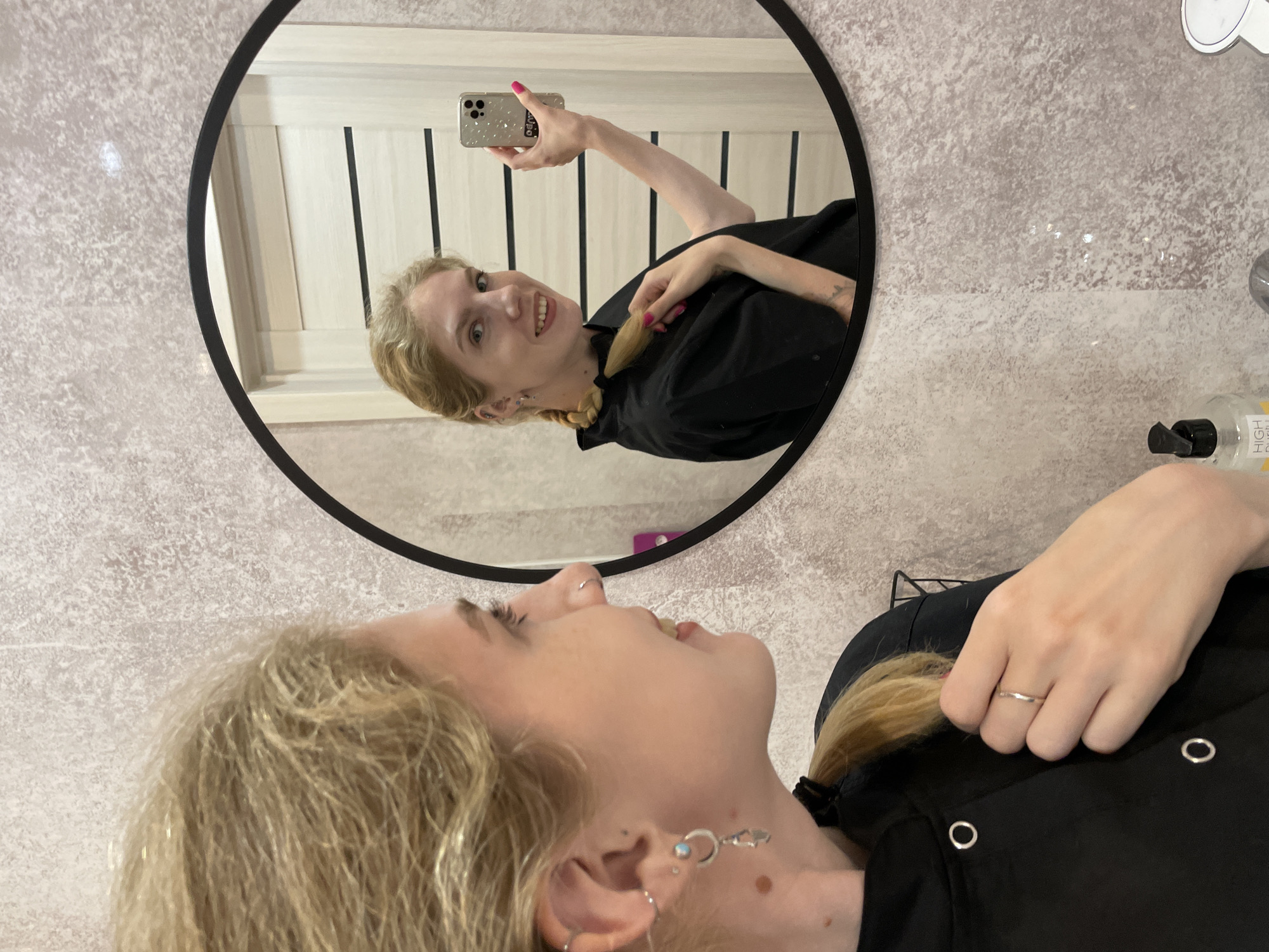 A young, blond woman is taking a selfie in a round mirror. The mirror reflects her holding a smartphone in her right hand, with a pink manicure, pointing the camera towards her face. She is smiling in the reflection. The reflection also shows a partially visible white door with vertical panels.
The woman in the foreground, seemingly the same person, is oriented at a different angle, revealing her profile. She has blond hair, a nose ring, and multiple earrings on her left ear. She is wearing a black shirt. A ring is visible on her left hand.
The background is a light gray, textured wall. It is unclear from the image if the location is in Izhevsk, Russia.