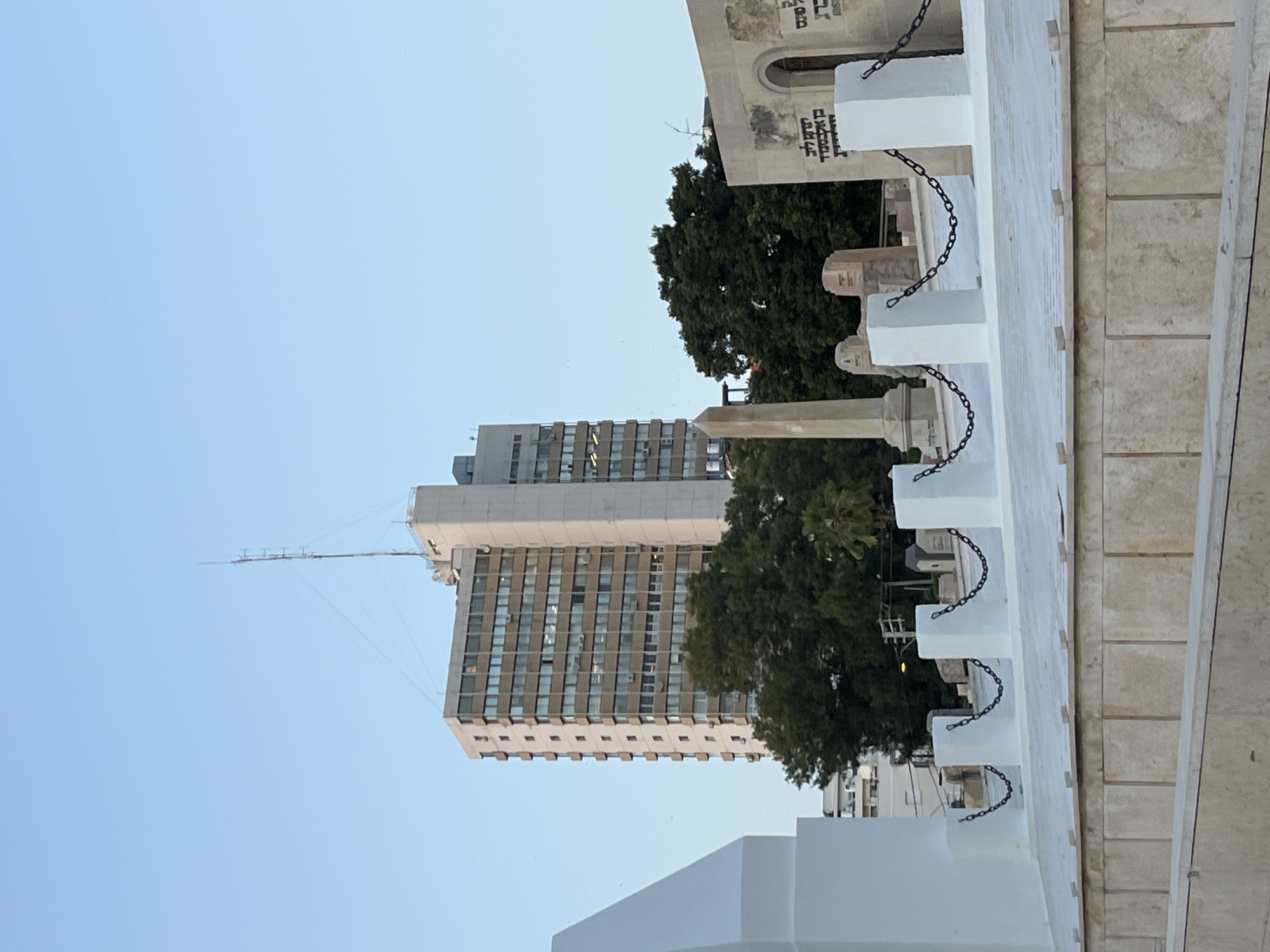 The image captures a view in Tel Aviv, Israel. A tall, rectangular building stands prominently in the background, its facade marked by rows of windows and a thin antenna extending from its top. The sky is a clear, light blue. In the foreground, a white structure with repeating architectural elements and chains is visible. A large tree is positioned between the building and the white structure. To the right, there is a pale stone wall. The scene gives the impression of a cemetery or memorial site.