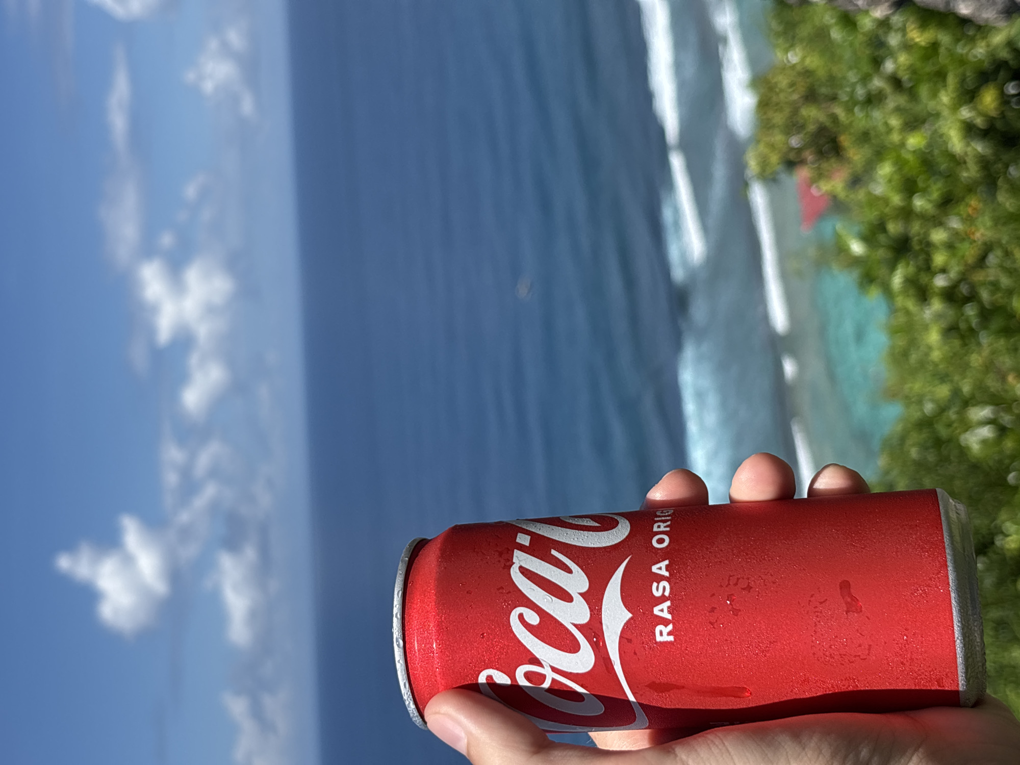 A close-up shot features a hand holding a red can of Coca-Cola, with condensation visible on its surface. The background displays a scenic view of the ocean, the horizon, and the sky with scattered clouds. The water appears blue and turquoise. The scene is at Ungasan, Indonesia. Green vegetation is visible to the right, suggesting a coastal cliffside location.