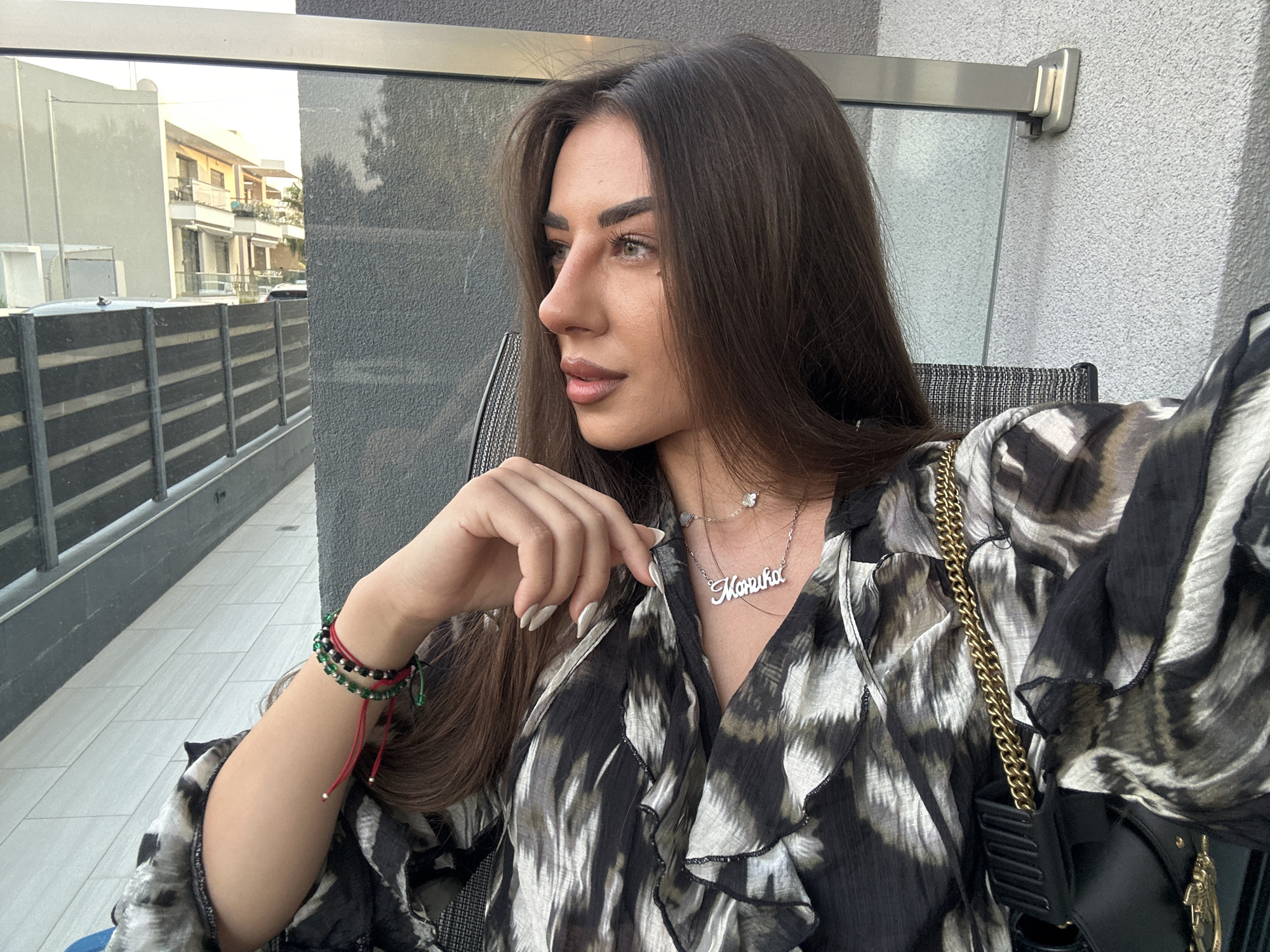A woman with long, dark brown hair is sitting outside in a modern, outdoor setting. She is wearing a black and white patterned blouse and a silver necklace with a nameplate that reads "Marika". She has bracelets on her left wrist, including red, black, and green beads. Her right hand is resting on her chest, near her necklace. The background shows a reflective surface, likely a glass railing or window, which reflects buildings and trees. The environment appears to be a patio or balcony area in the city of Ofrynio Beach, Greece, with a modern building and outdoor fencing visible.