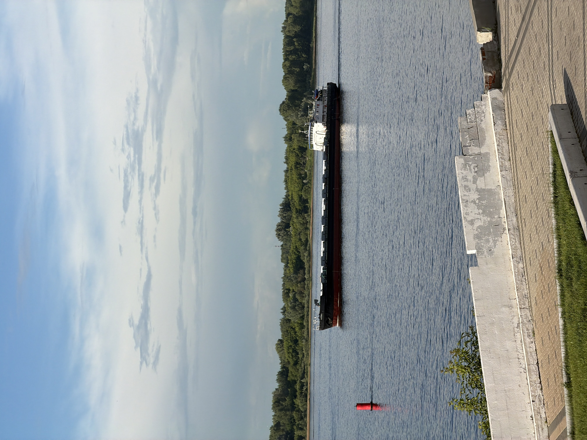 A barge with a white superstructure and a black hull, red waterline, is sailing on a wide, calm river. The barge is loaded with rectangular cargo covered with tarpaulin. To the left of the barge, a red buoy is visible in the water. In the background, a dense green forest lines the opposite bank of the river. The sky is partially cloudy with a mix of blue sky and white clouds. In the foreground, there is a paved walkway and a white concrete barrier wall. The overall scene depicts a sunny day with moderate cloud cover on a river in the city of Nizhny Novgorod, Russia, likely during daytime.