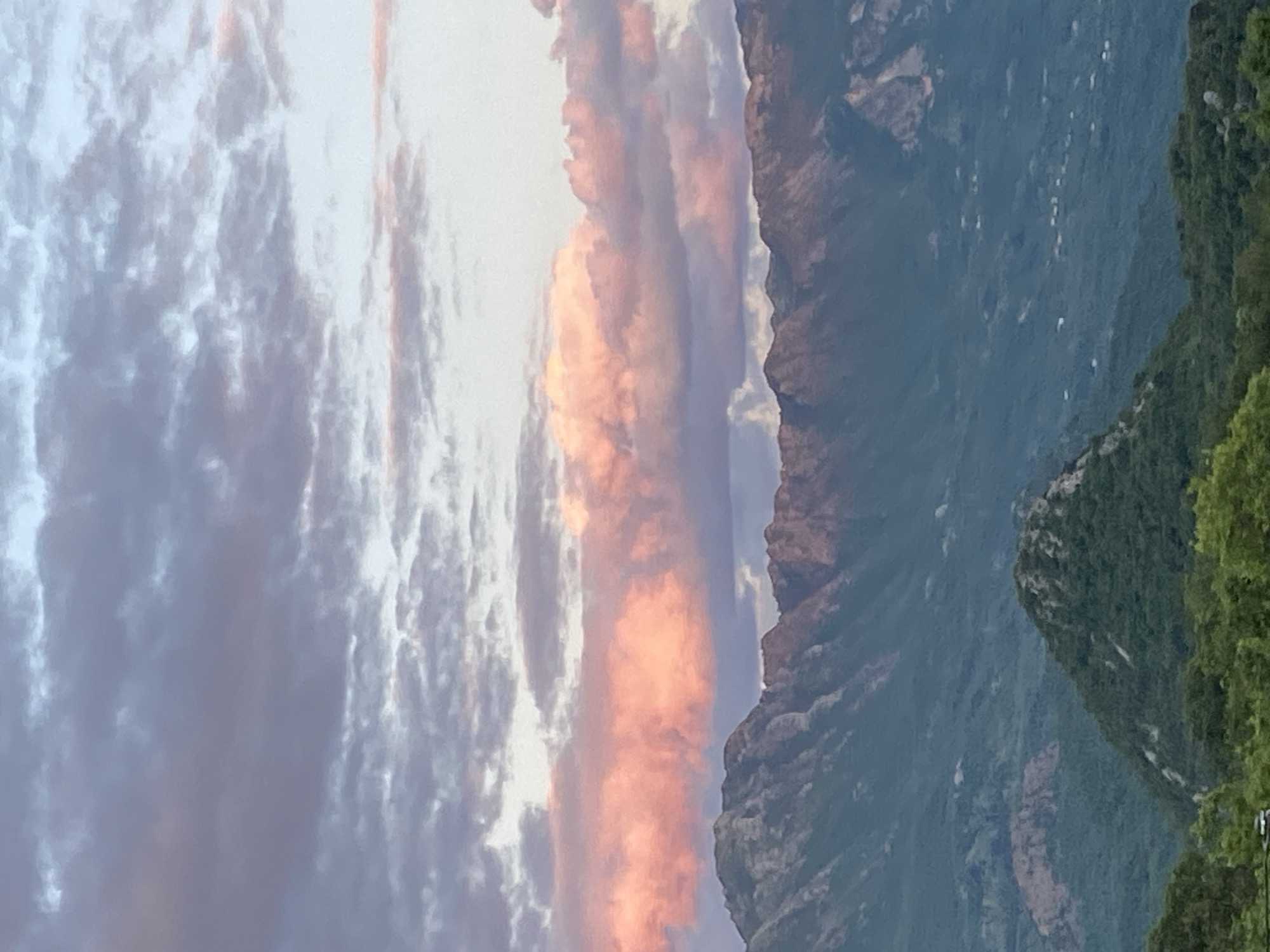 This vertical landscape image captures a dramatic mountain scene under a colorful sky, likely at sunrise or sunset. The scene is dominated by a majestic mountain range that stretches across the middle ground, featuring rugged, dark green slopes covered in dense vegetation, with hints of grey rock. Multiple layers of mountains recede into the distance, creating a sense of depth and vastness.
Above the mountains, the sky is filled with clouds. A prominent horizontal band of clouds, roughly in the center, glows with warm orange and pink hues, strongly suggesting either the first light of dawn or the last light of dusk breaking through. Above and below this luminous band, the clouds are a softer blend of grey, white, and subtle purplish tones, indicating varying light conditions and cloud density.
To the right side of the image, closer to the viewer, is a steep, heavily forested slope densely covered with vibrant green foliage, appearing as a dark, textured mass that frames the more distant mountains and the sky. No human activity, objects, or visible text are present. The image conveys a serene and awe-inspiring natural vista, characteristic of a mountainous region like Sotonići, Montenegro, during a golden hour.