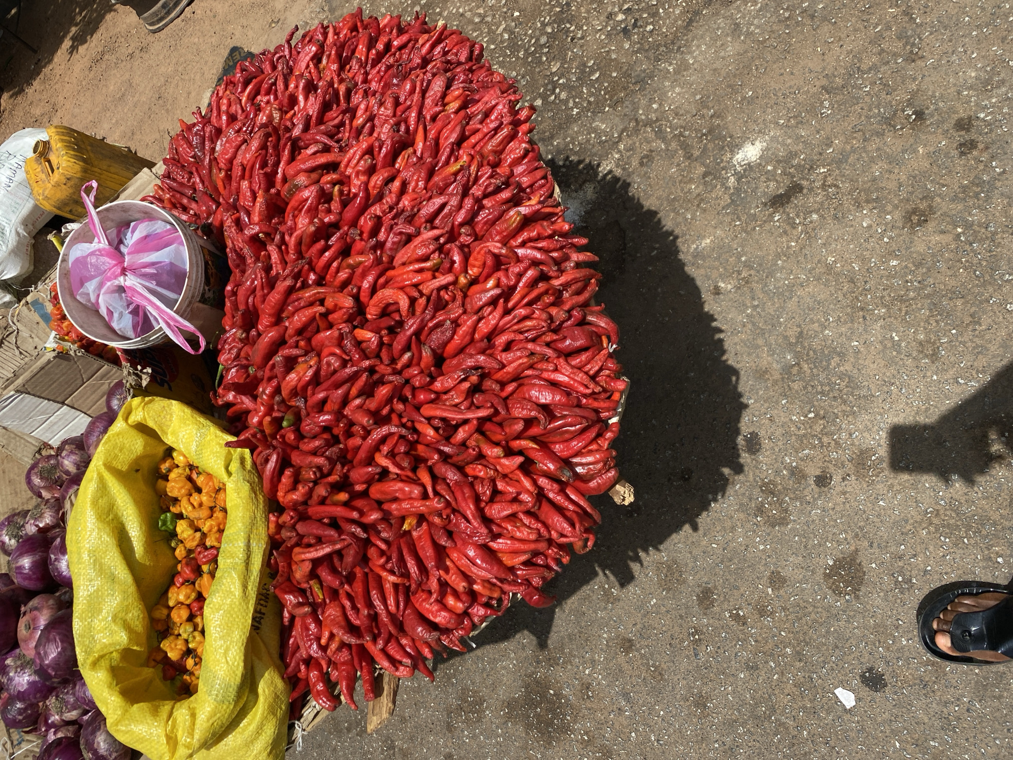 This image, taken in Bukuru, Nigeria, shows a street-level vendor's wares arranged on the ground. A large pile of red chili peppers dominates the center, casting a distinct shadow on the concrete. To the left of the peppers, there's a yellow sack filled with orange and yellow peppers, along with a pile of purple onions. Near the peppers is a white bucket holding a plastic bag with pink and white stripes. A yellow plastic container and cardboard boxes are visible behind the bucket. In the lower right corner, a foot wearing a black sandal is visible on the concrete pavement. The overall scene depicts a typical market setting in Bukuru, Nigeria.