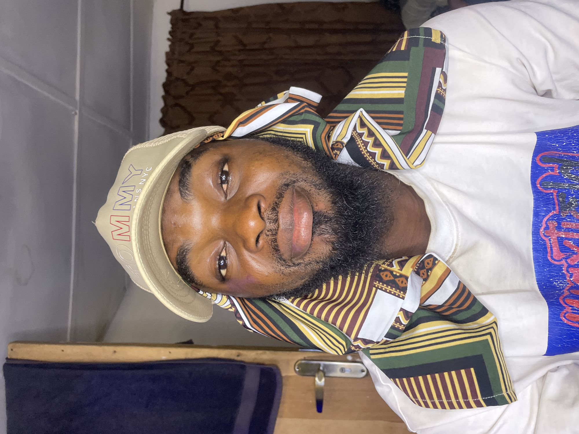 A man with dark skin and a full beard is pictured from a close, angled perspective. He is wearing a light-colored baseball cap with the word "TOMMY" embroidered on it. The man has a patterned scarf wrapped around his head and neck, featuring stripes and geometric shapes in green, yellow, brown, and white. He is wearing a white t-shirt. The background is slightly blurred, but hints of a curtain or draped fabric can be seen. The photo appears to be taken indoors. There are no visible indicators to confirm that it was taken in Jos, Nigeria.